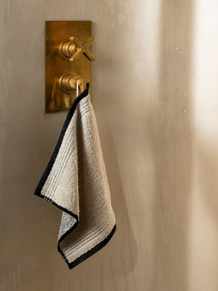 Handtuch (hellbeige mit dunklem Rand) hängt an goldfarbener Messingarmatur an einer hellen, leicht feuchten Badezimmerwand; textile Struktur sichtbar, dezente seitliche Beleuchtung.