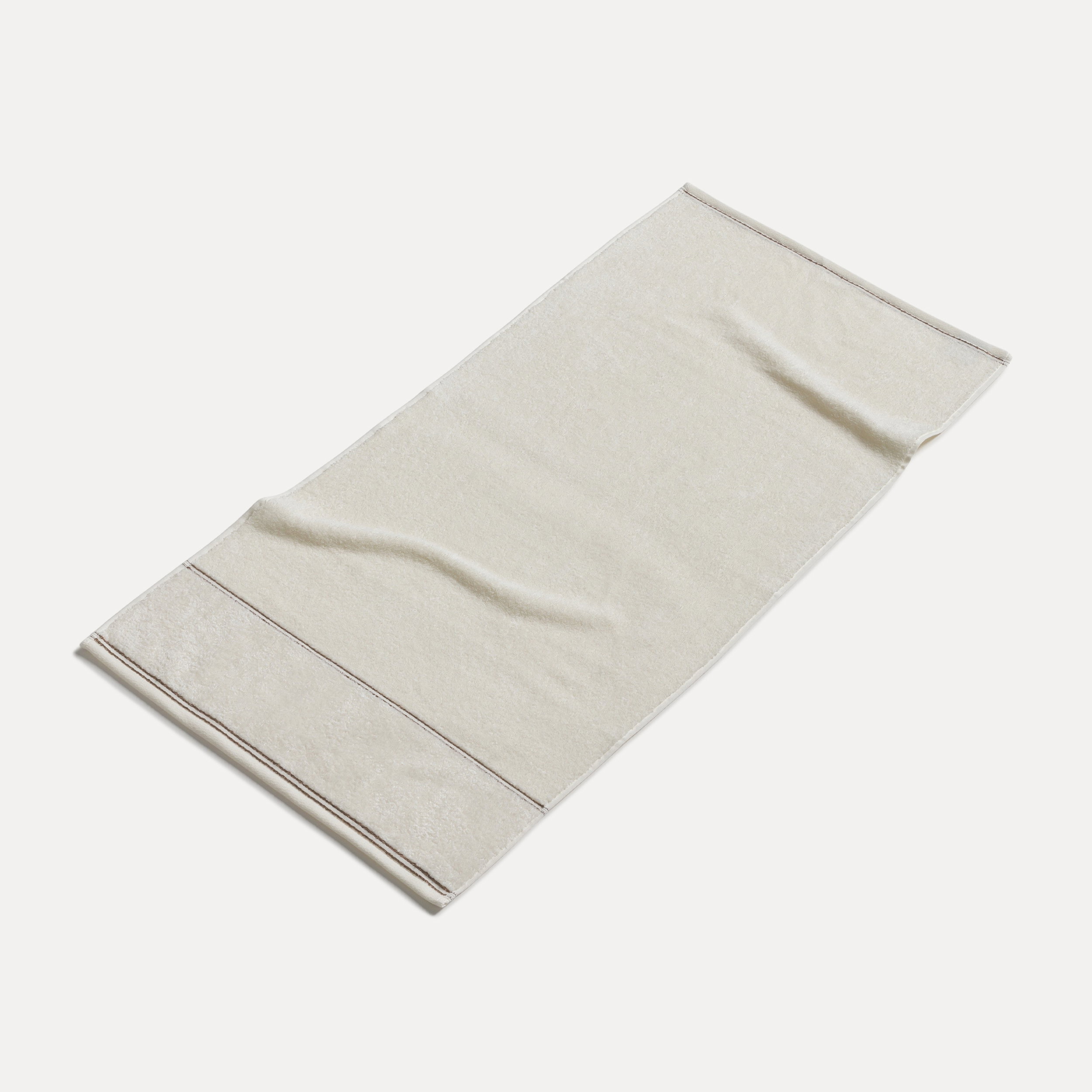 Handtuch liegt ausgebreitet und leicht gewellt auf neutralem, hellem Untergrund; naturfarben mit feiner Textur und dezenter Bordüre am Rand.