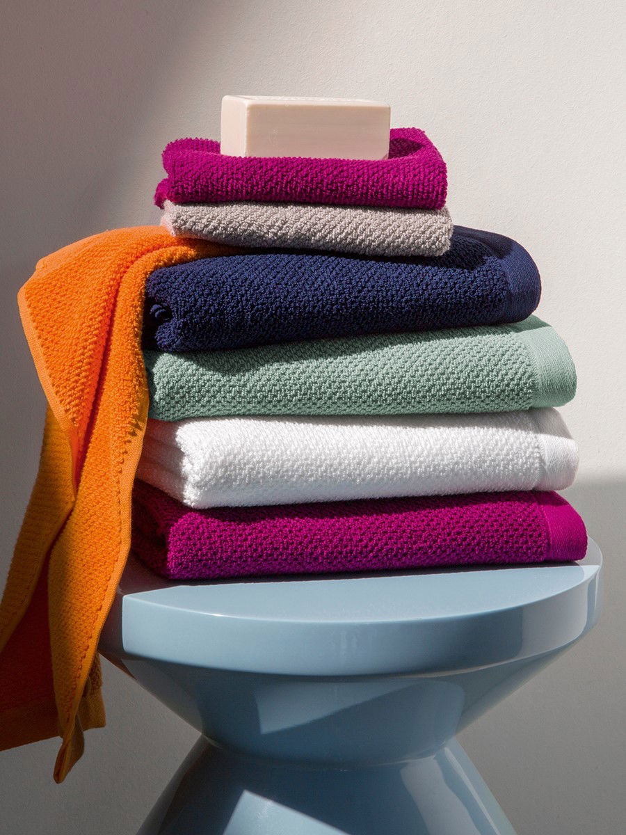 Mehrere farbige Möve-Handtücher (orange, pink, weiß, navy, mint) sind zu einem Stapel gefaltet, mit einem Seifenstück obenauf; der Stapel steht auf einem hellblauen Hocker vor neutraler Wand.