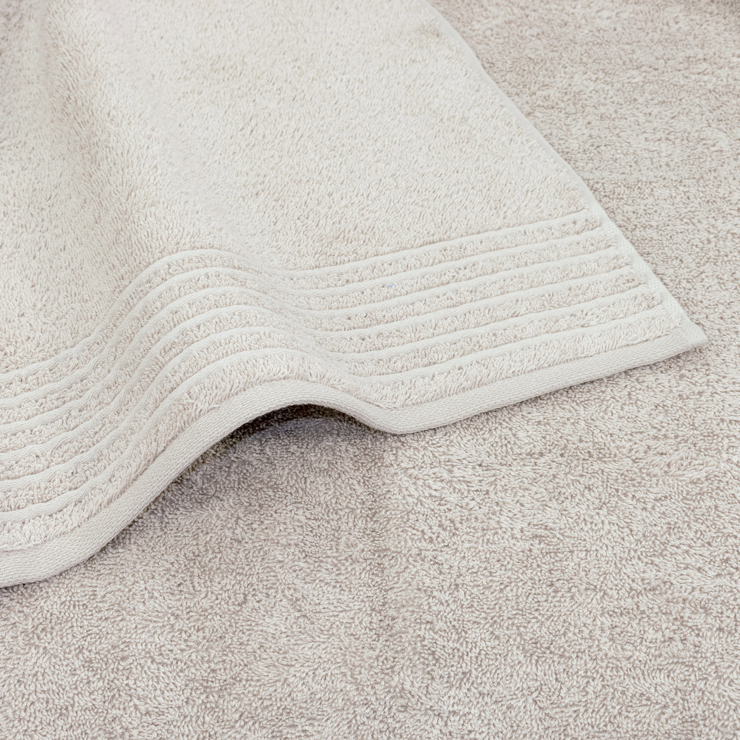 A light beige towel lies flat, one corner slightly turned up; a woven border with fine stripes is visible against a matching towel background in the same shade.