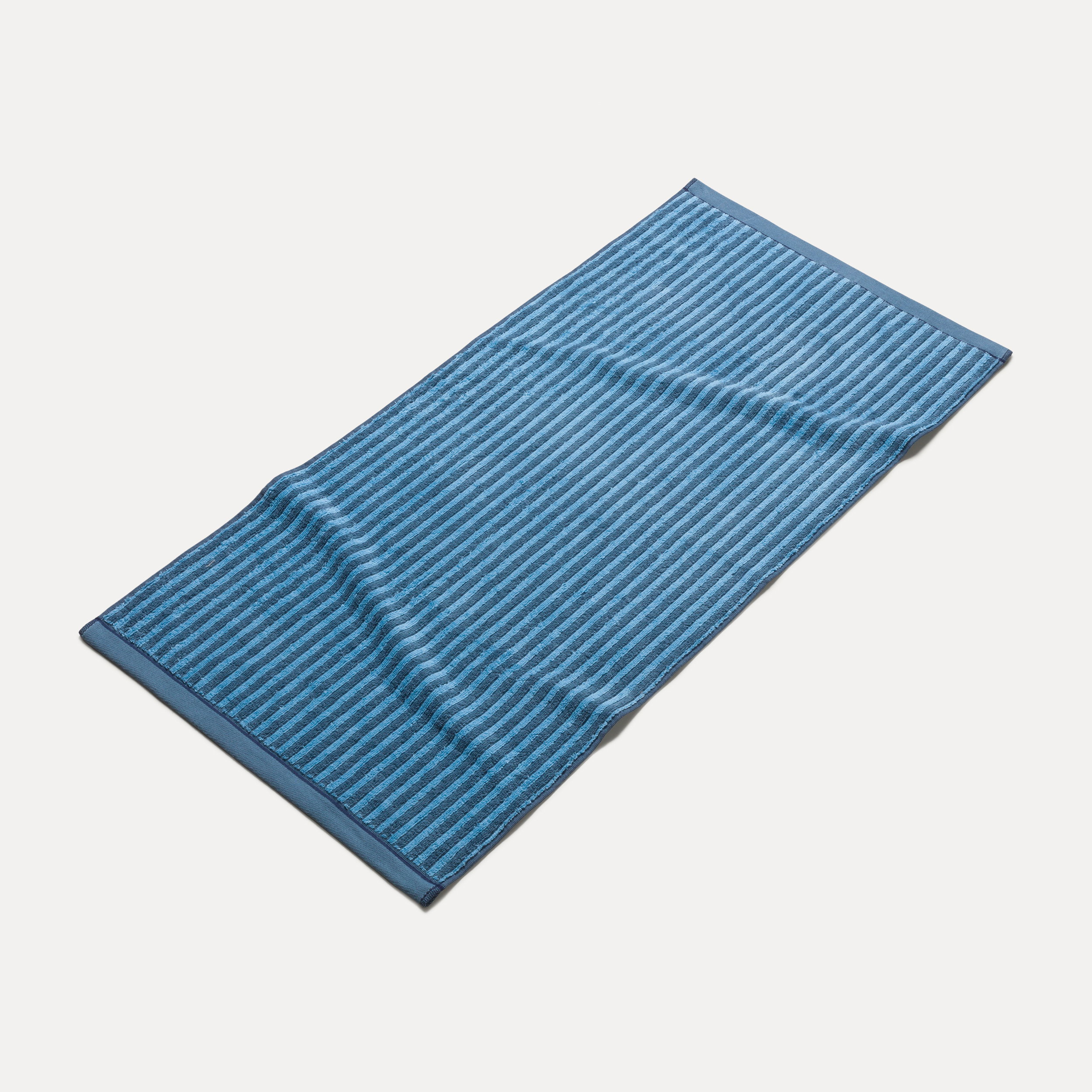 Ein blau gestreiftes Badetuch liegt flach ausgebreitet, leicht zerknittert, auf einem neutralen hellen Hintergrund.