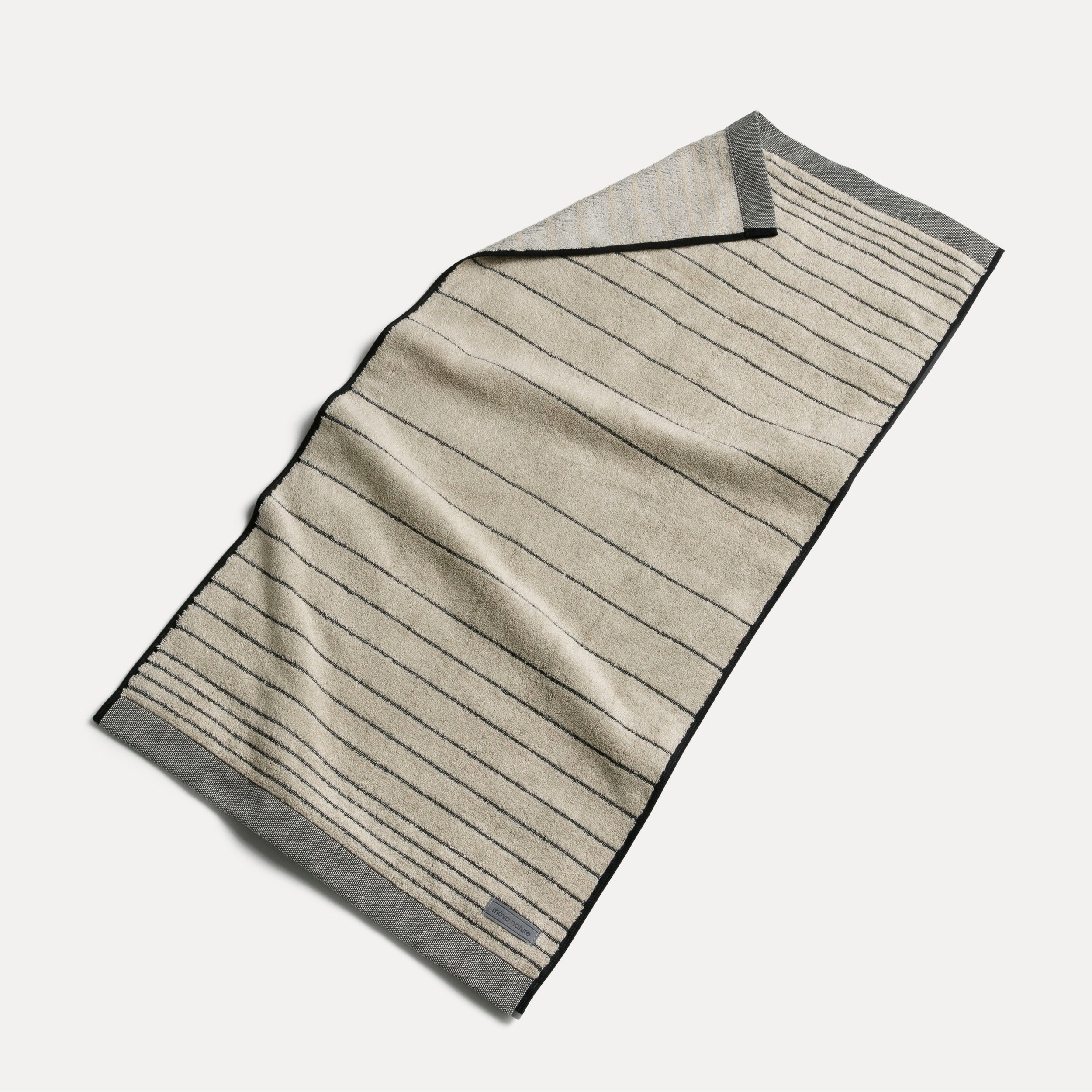 Bath towel, lying slightly spread out, shows fine horizontal stripes on a light beige background with a dark gray border; flat, neutral background, textile texture and hem visible.