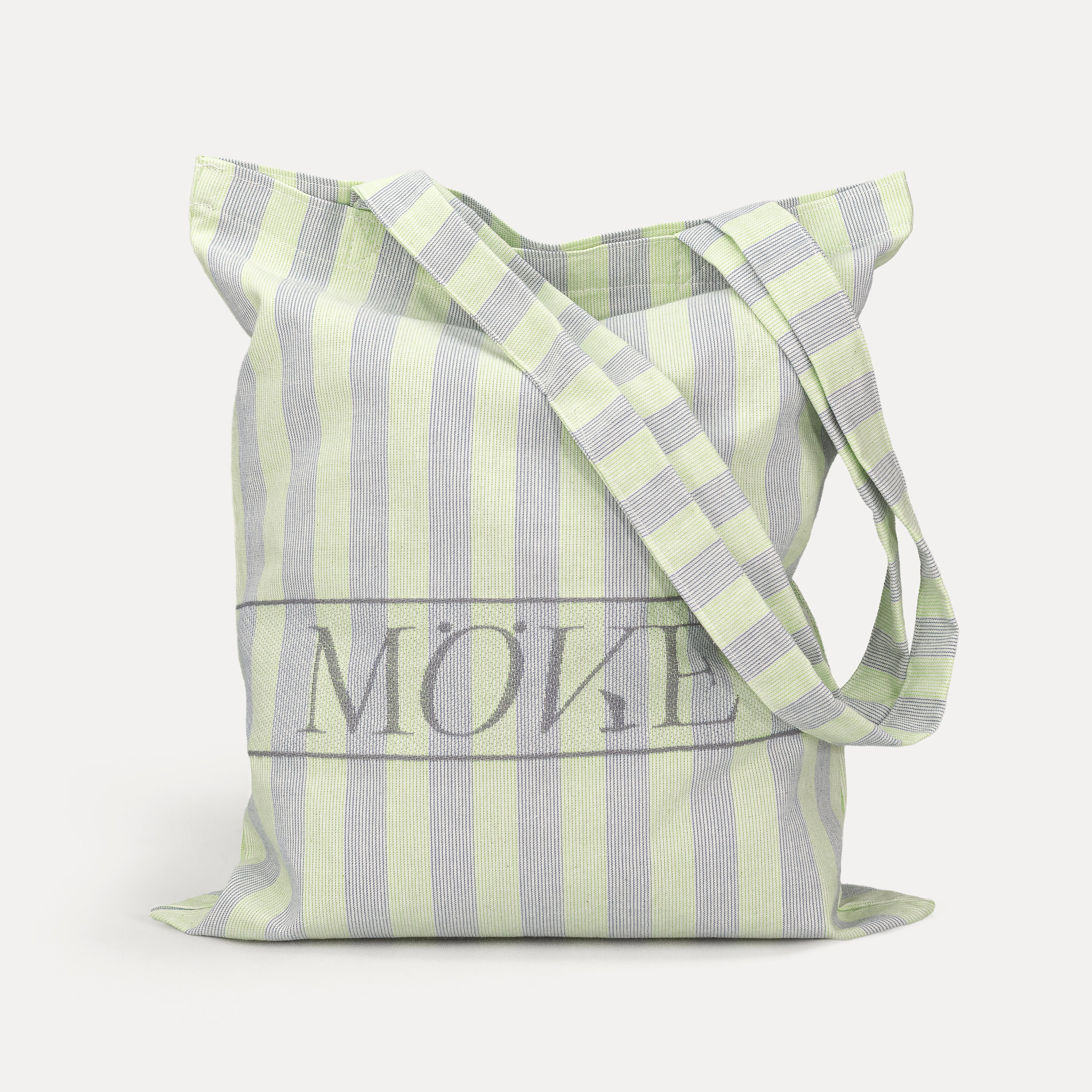 Fabric tote bag with wide vertical light green-gray stripes and a large "MÖVE" logo, hanging loosely from a long shoulder strap, isolated against a neutral, light gray background.