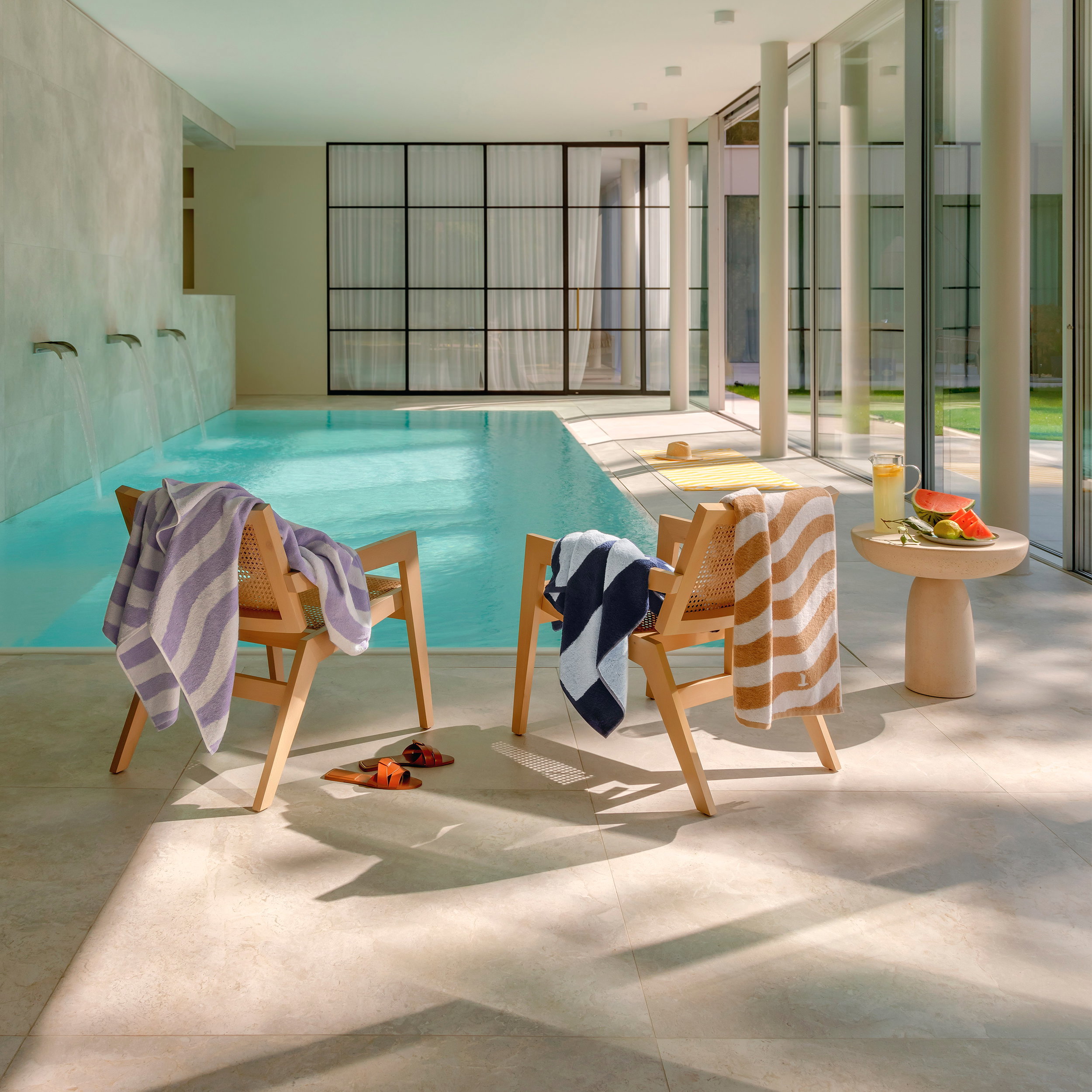 Striped towels hang over two wooden chairs; next to them is a small side table with a drink and fruit. Context: bright, modern indoor swimming pool with large windows.