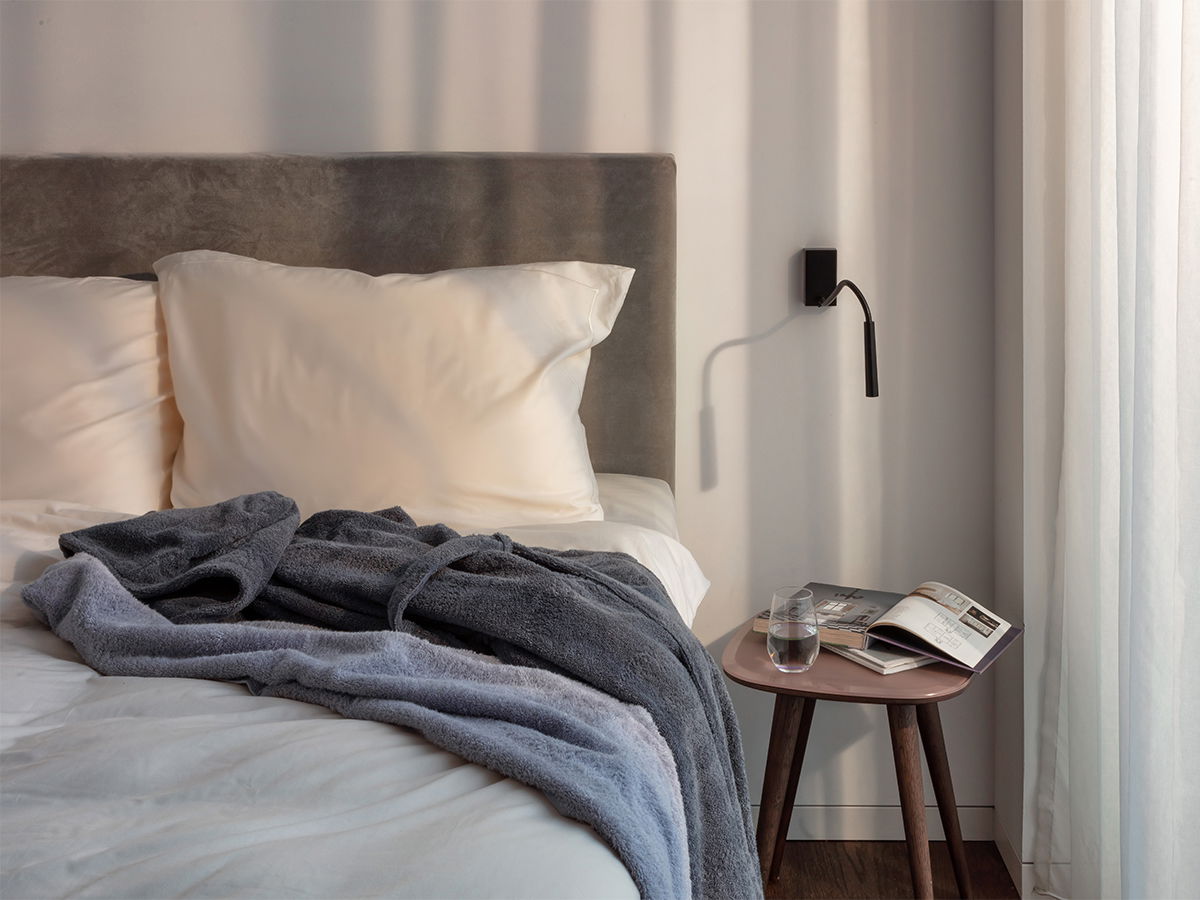Graues Frottee-Handtuch liegt lässig zerwühlt auf einem weißen Bett; Kissen und gepolstertes Kopfteil, kleiner Nachttisch mit Glas und offenem Magazin, heller Vorhang im Hintergrund.