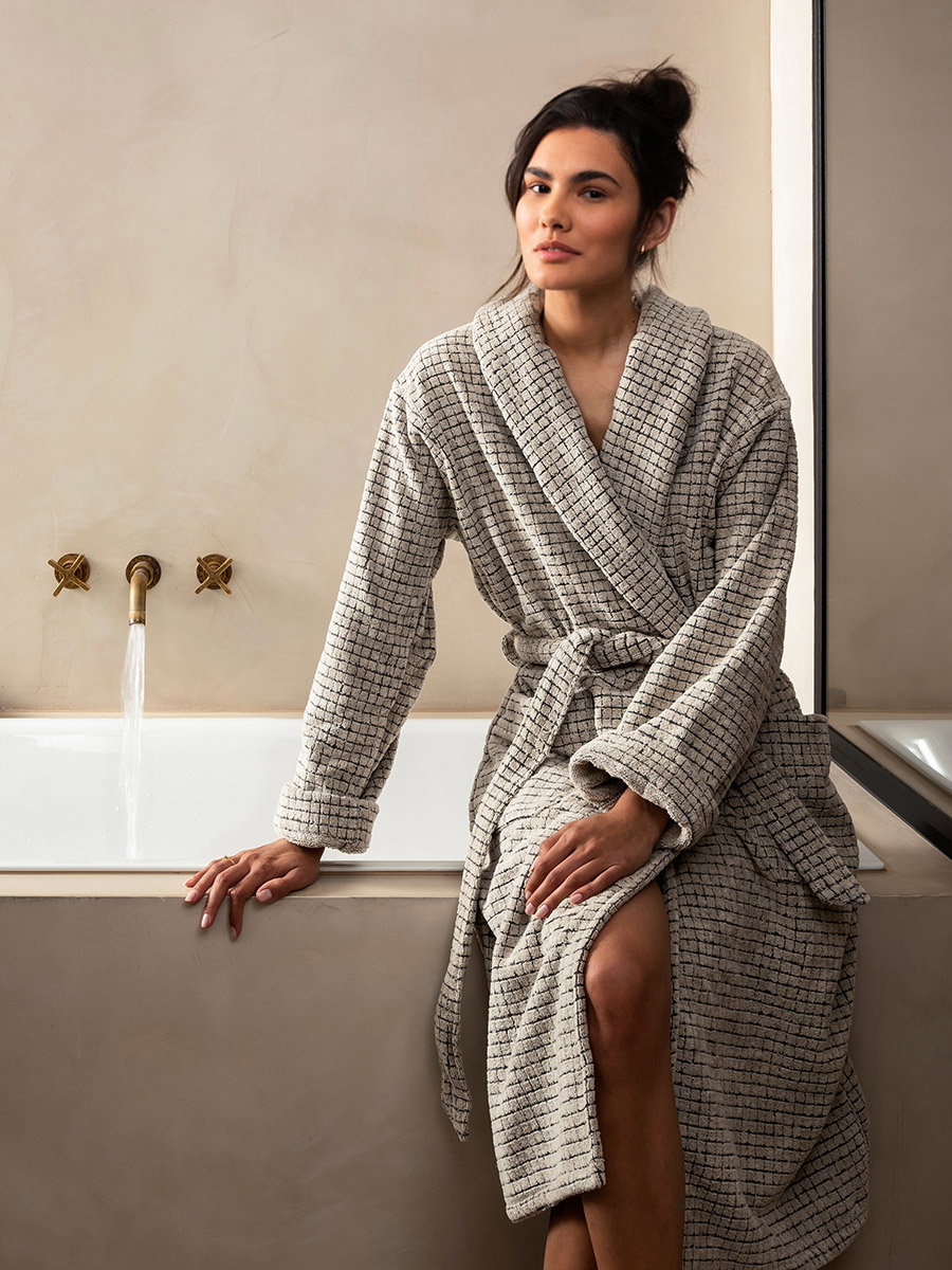 Waffle-weave bathrobe (gray)—rested casually on the edge of the bathtub—in a simple bathroom with a cream-colored wall, built-in bathtub, two bronze-colored faucets, and natural light.