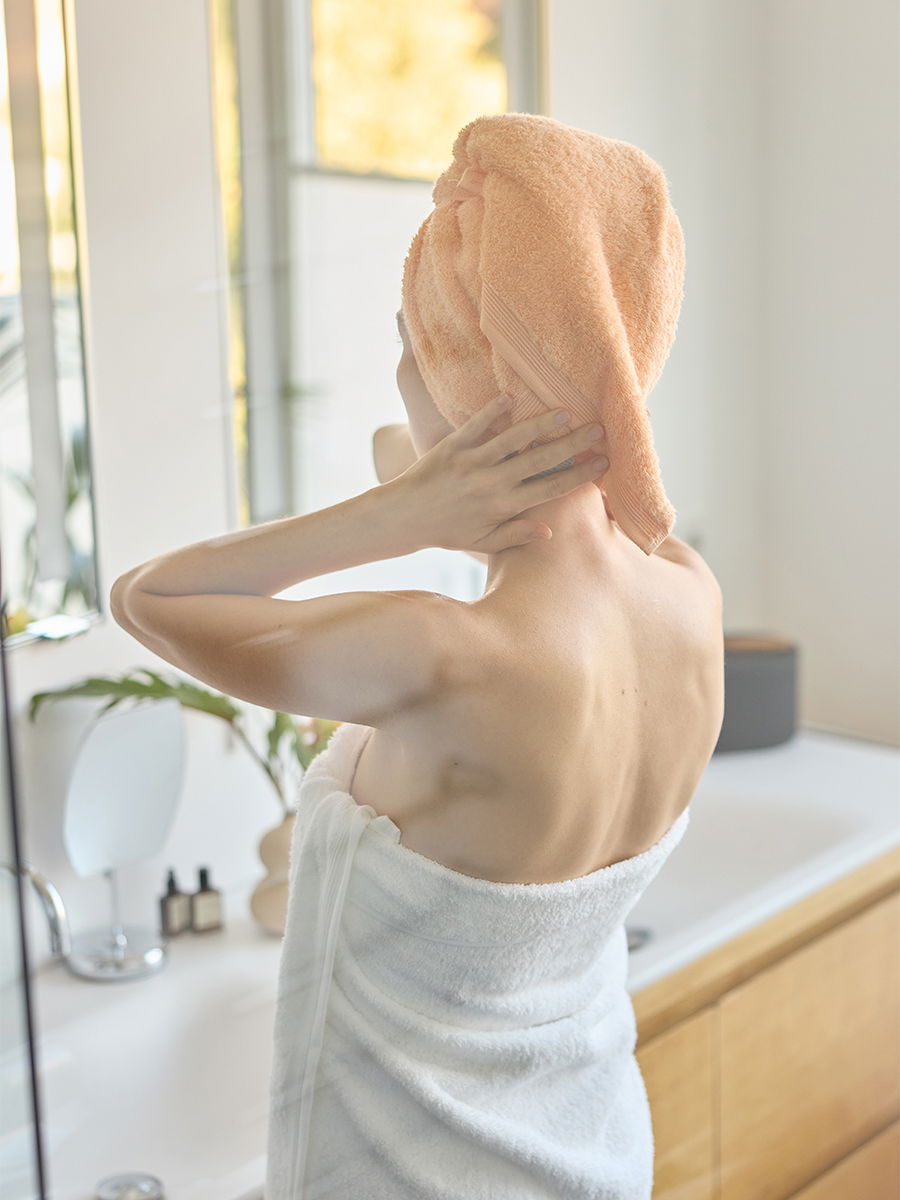 Orangenes Haartuch um den Kopf einer Person, die es mit beiden Händen justiert; sie trägt ein weißes Badetuch und steht vor einem hellen Waschbecken im Badezimmer.