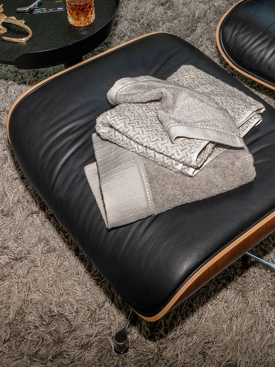 Mehrere beige-graue Möve-Handtücher mit strukturierter Webung liegen gefaltet auf einem schwarzen Lederhocker; Kontext: dichter heller Teppich, daneben runder Beistelltisch mit Glas, Schlüssel und Dekor.
