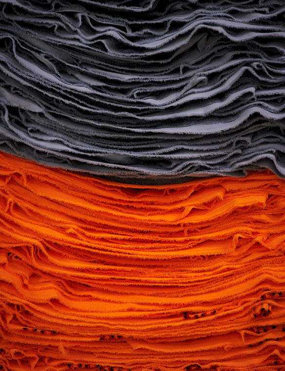 Handtücher – graue und orangefarbene Frottee-Lagen liegen in dichten Stapeln übereinander; ausgeprägte Kanten und Fasern sichtbar; Nahaufnahme in neutralem Studio-/Lagerkontext.