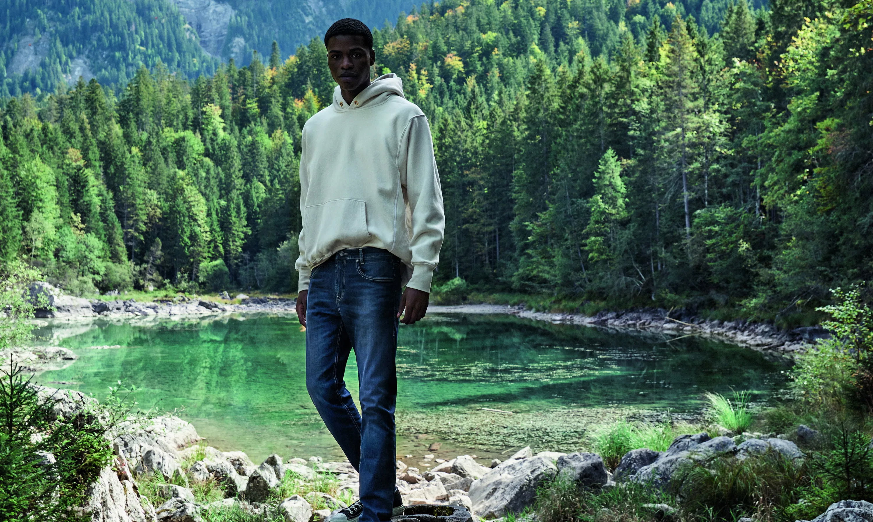 Person in hellem Hoodie und Jeans steht am Ufer; klarer grüner Bergsee im Vordergrund, dichter Nadelwald und felsiges Ufer bilden den natürlichen Hintergrund.