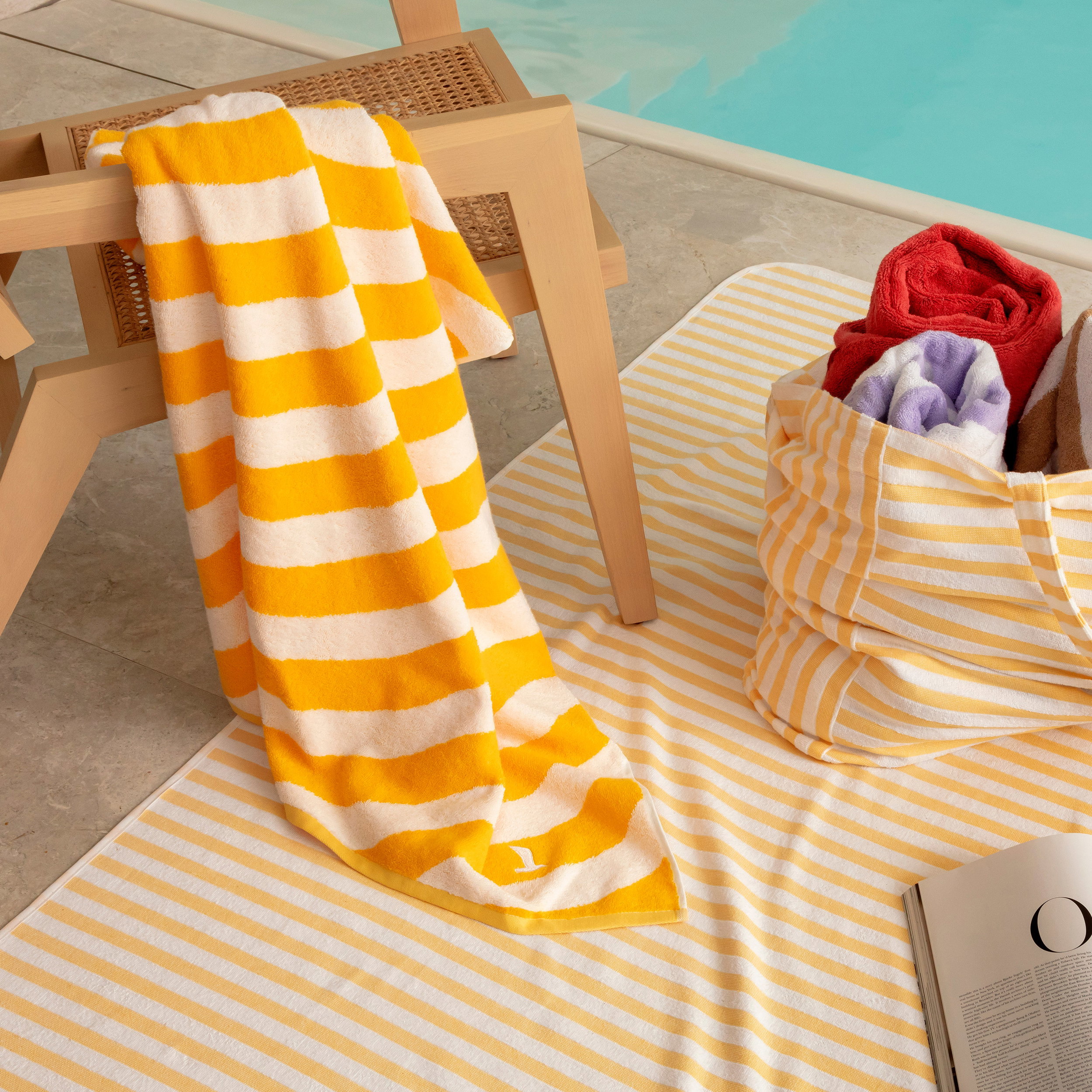 A striped yellow-and-white towel hangs over a wooden chair; a striped mat and a fabric basket with rolled-up towels next to it; pool area with water in the background.