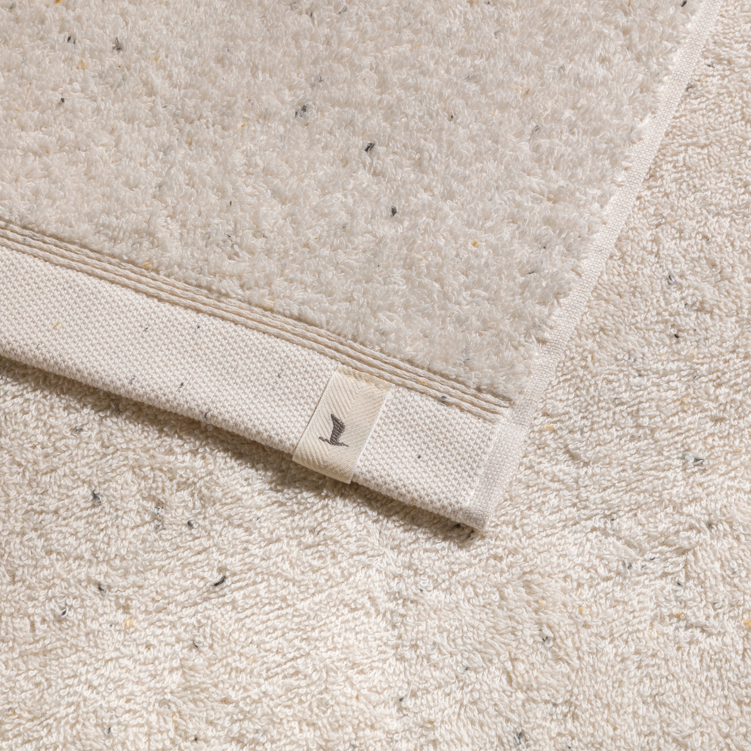 A beige terry towel with a woven edge and Möve label lies on a matching, lightly speckled bath mat; a close-up shows the soft texture and a light, neutral-colored setting.