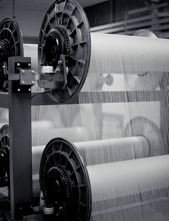 Große Spulen spannen feine Textilfäden; industrielle Webmaschine zeigt mehrere aufgewickelte Stoffbahnen im Fabrikumfeld.
