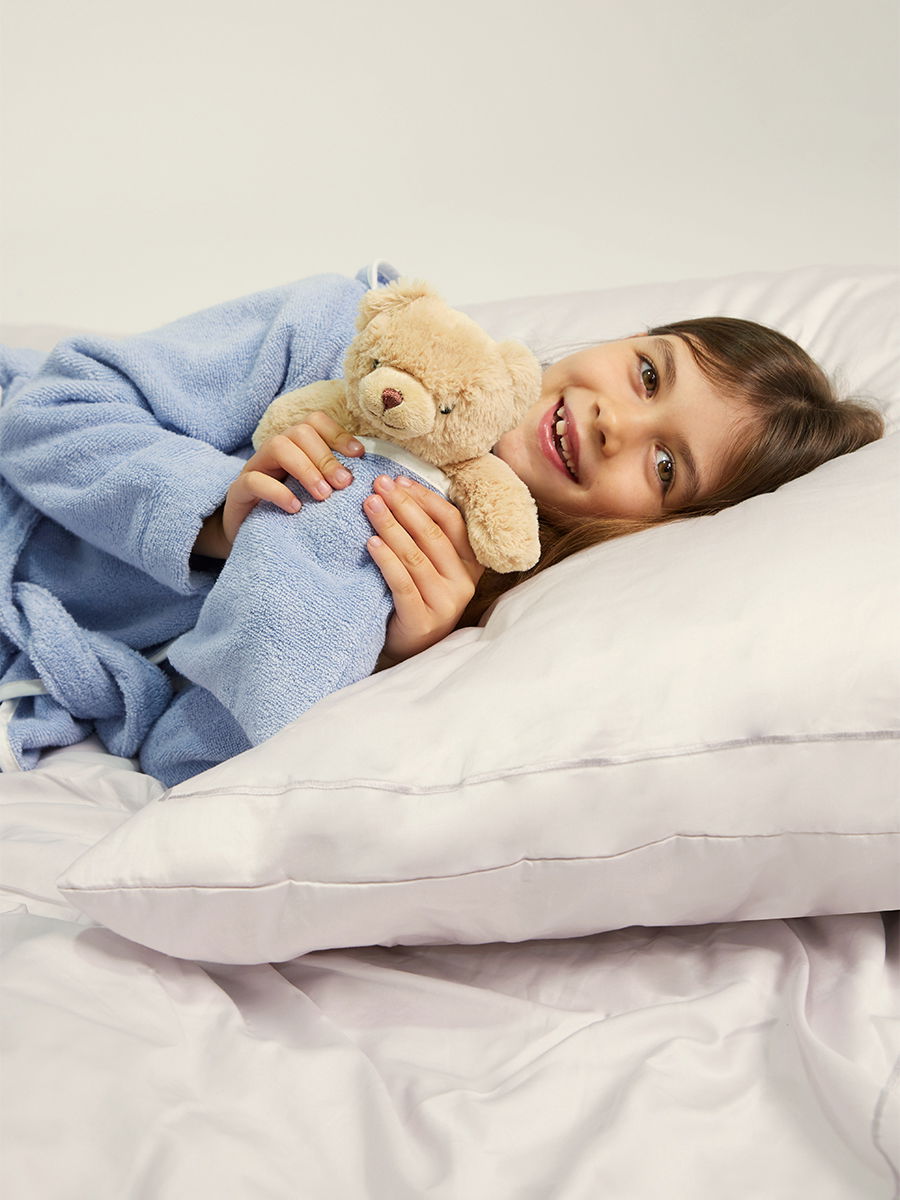 Child in a blue bathrobe lies smiling on white pillows, holding a small teddy bear; calm bedroom/studio setting with a neutral background.