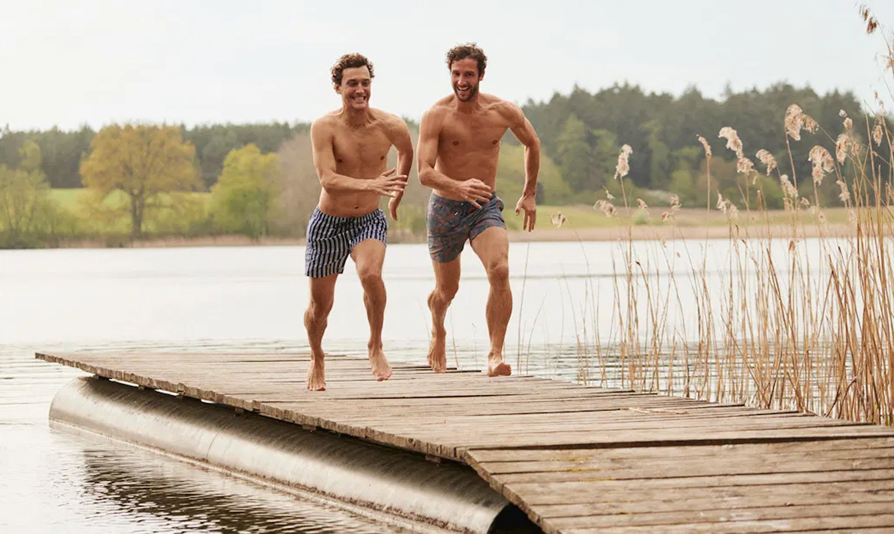 Zwei Männer in gestreiften bzw. gemusterten Badehosen laufen lachend über einen hölzernen Steg am See; Schilf und bewaldetes Ufer im Hintergrund.