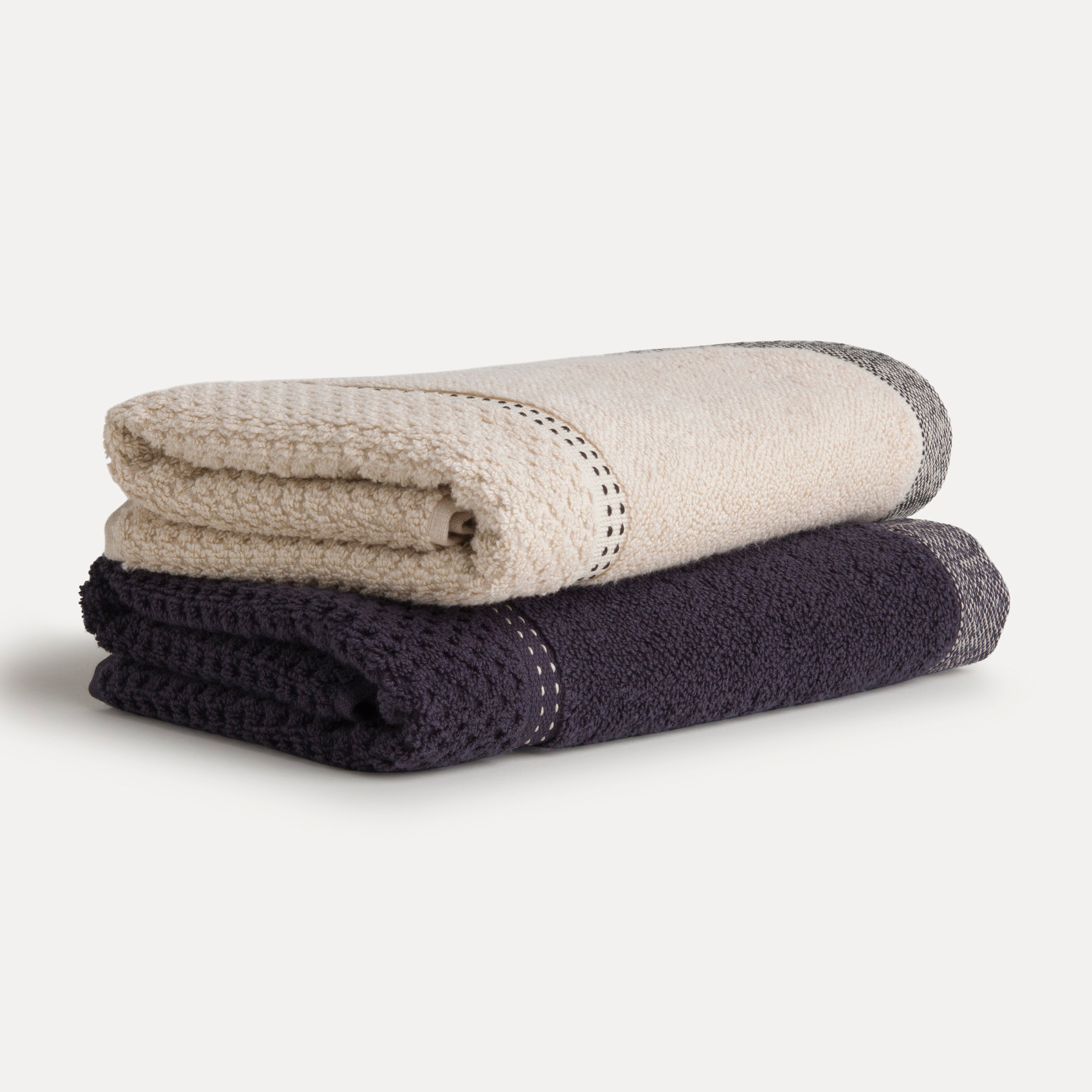 Zwei gefaltete Handtücher (oberes hellbeige mit grauem Rand, unteres dunkelgrau) liegen übereinander auf hellem einfarbigem Hintergrund; sichtbare Waffelstruktur und gestrickte Kanten.