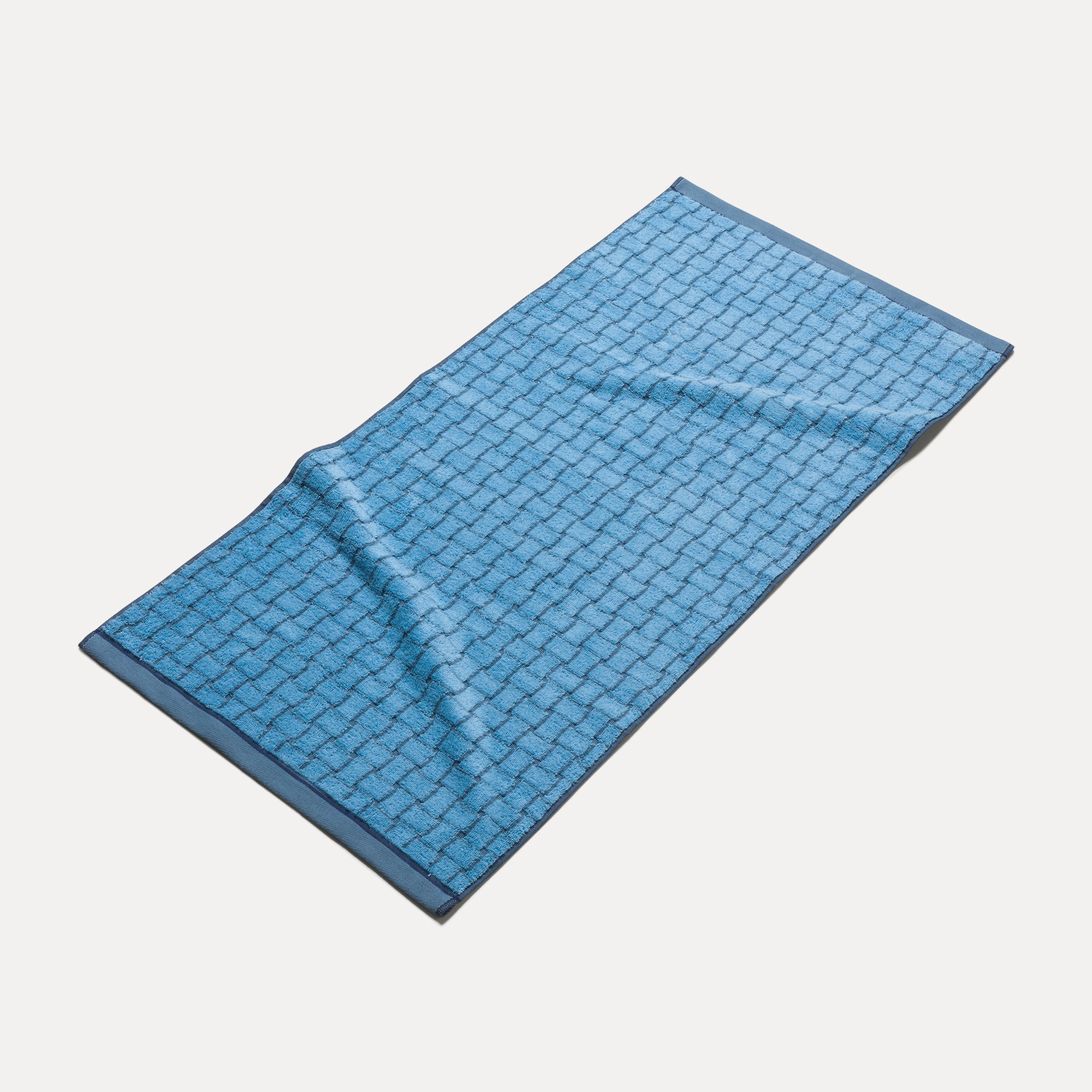 A blue towel lies spread out; it features a checkered textured pattern, slight wrinkles, and a darker border, photographed against a neutral, light background.
