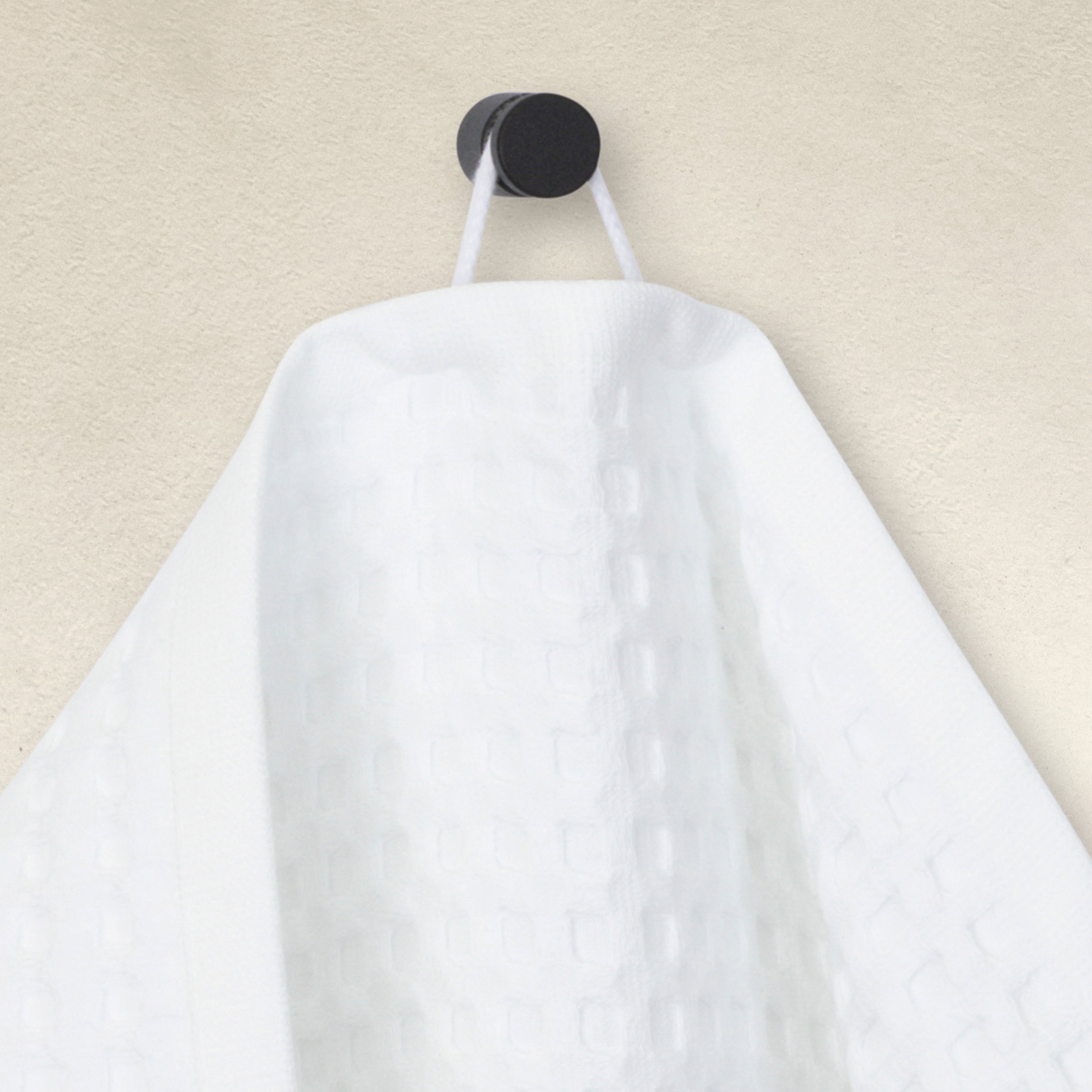 A white waffle-weave towel hangs on a round, dark wall hook; it falls in visible folds, showing a textured surface; context: light, minimalist wall surface.