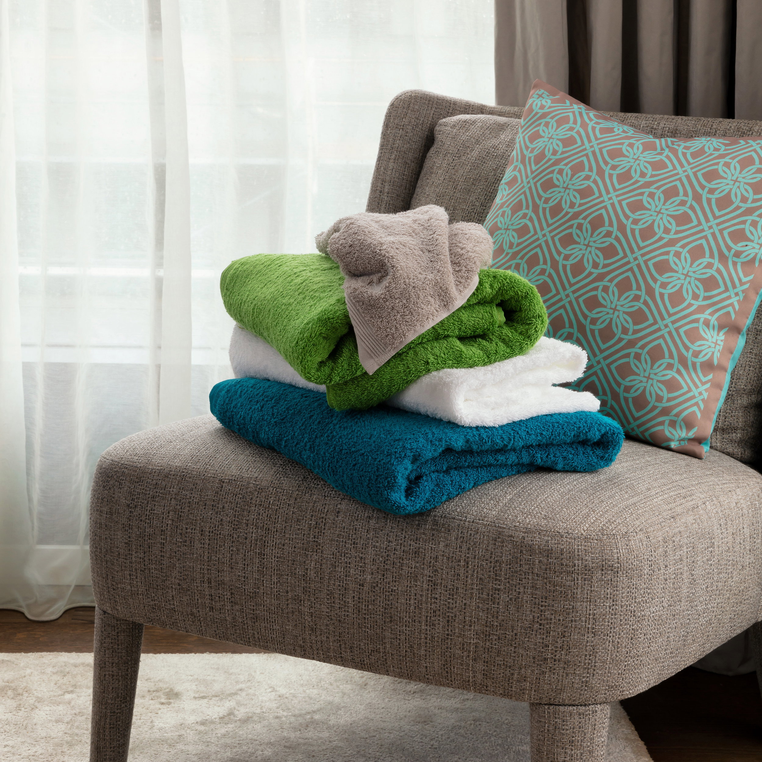 Three folded hand towels (turquoise, white, green) with a small beige hand towel on top lie on a gray armchair next to a patterned pillow and curtain.