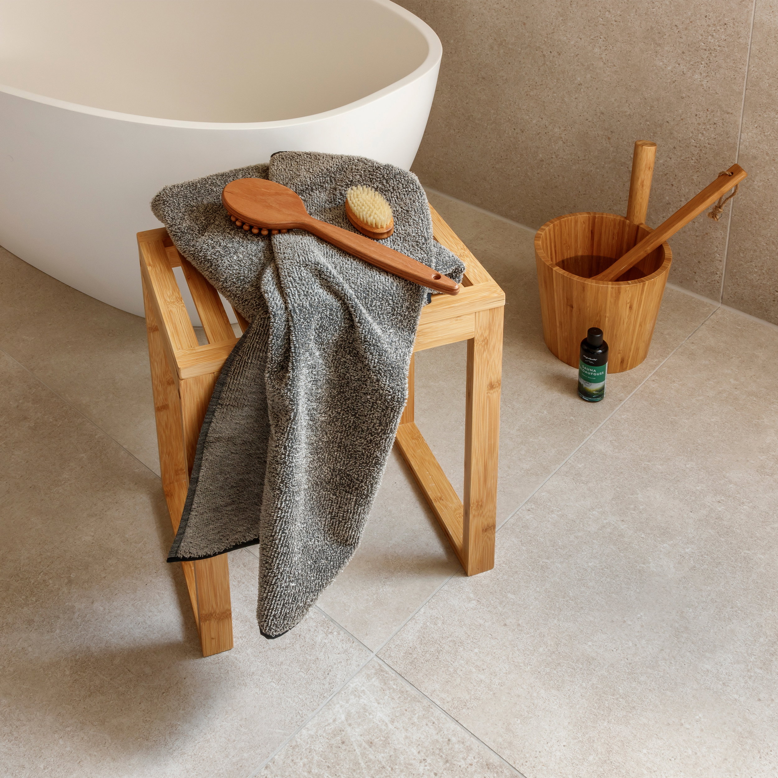 Graues Handtuch liegt auf einem kleinen Holz-Hocker; zwei Holzbürsten darauf. Neben der Badewanne ein Bambuseimer mit Kelle und eine Flasche auf sandfarbenen Fliesen.