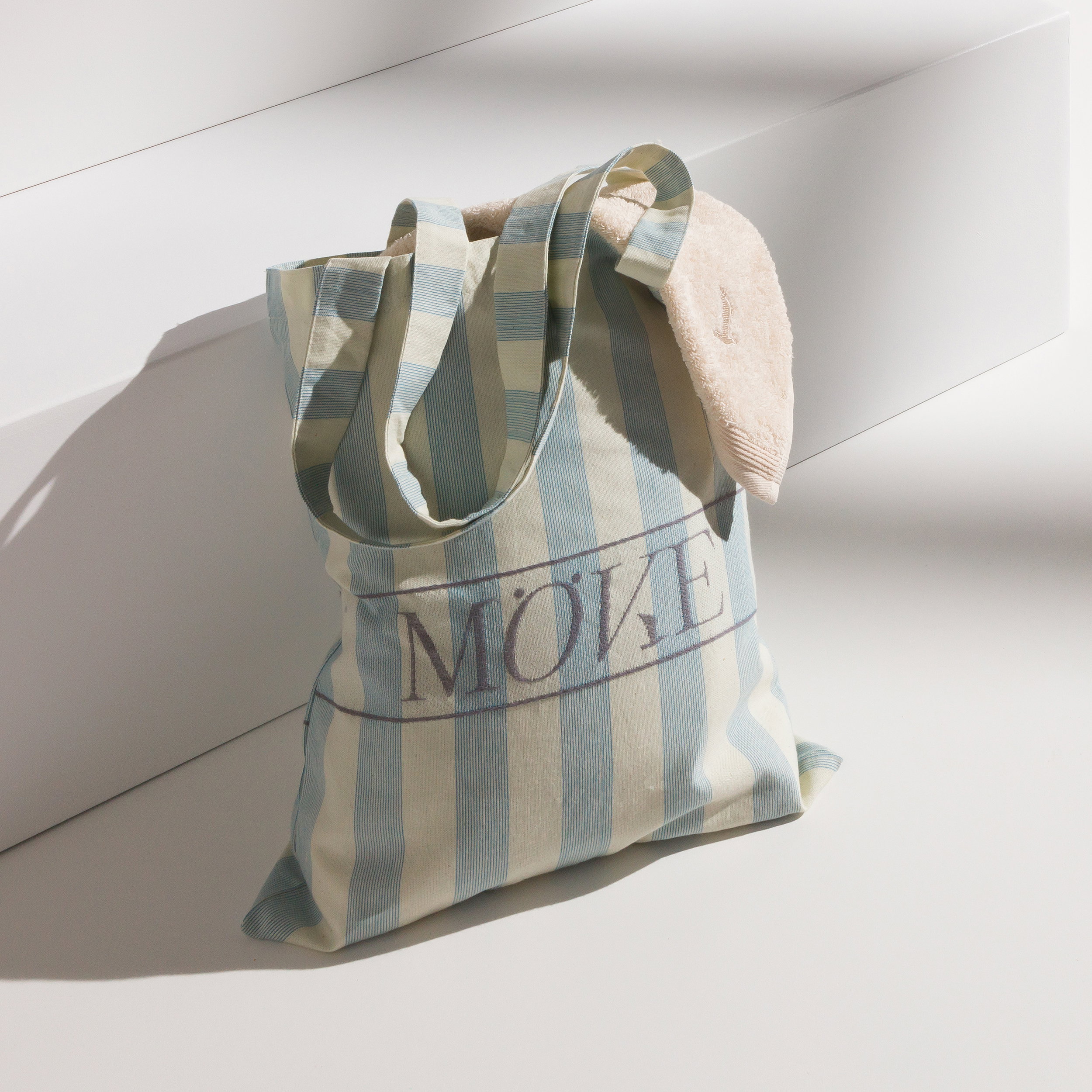 Striped fabric bag with "MÖVE" logo; a light-colored towel protrudes; bag rests on a white stair landing, soft sunlight highlights the material and shadows.
