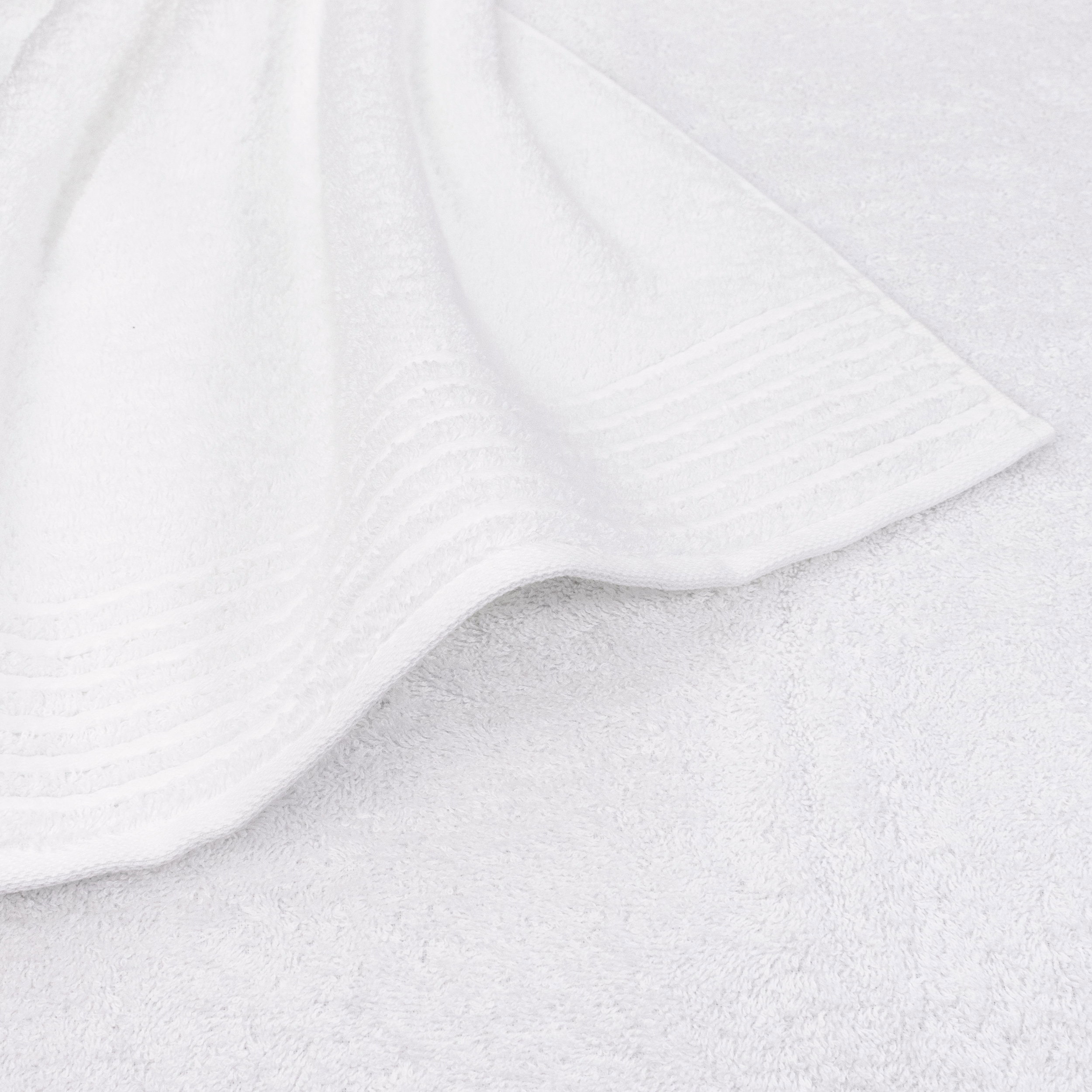 White towel, slightly turned up and curled at one corner, lies on another white towel; even texture, subtle striped trim, functional studio shot.