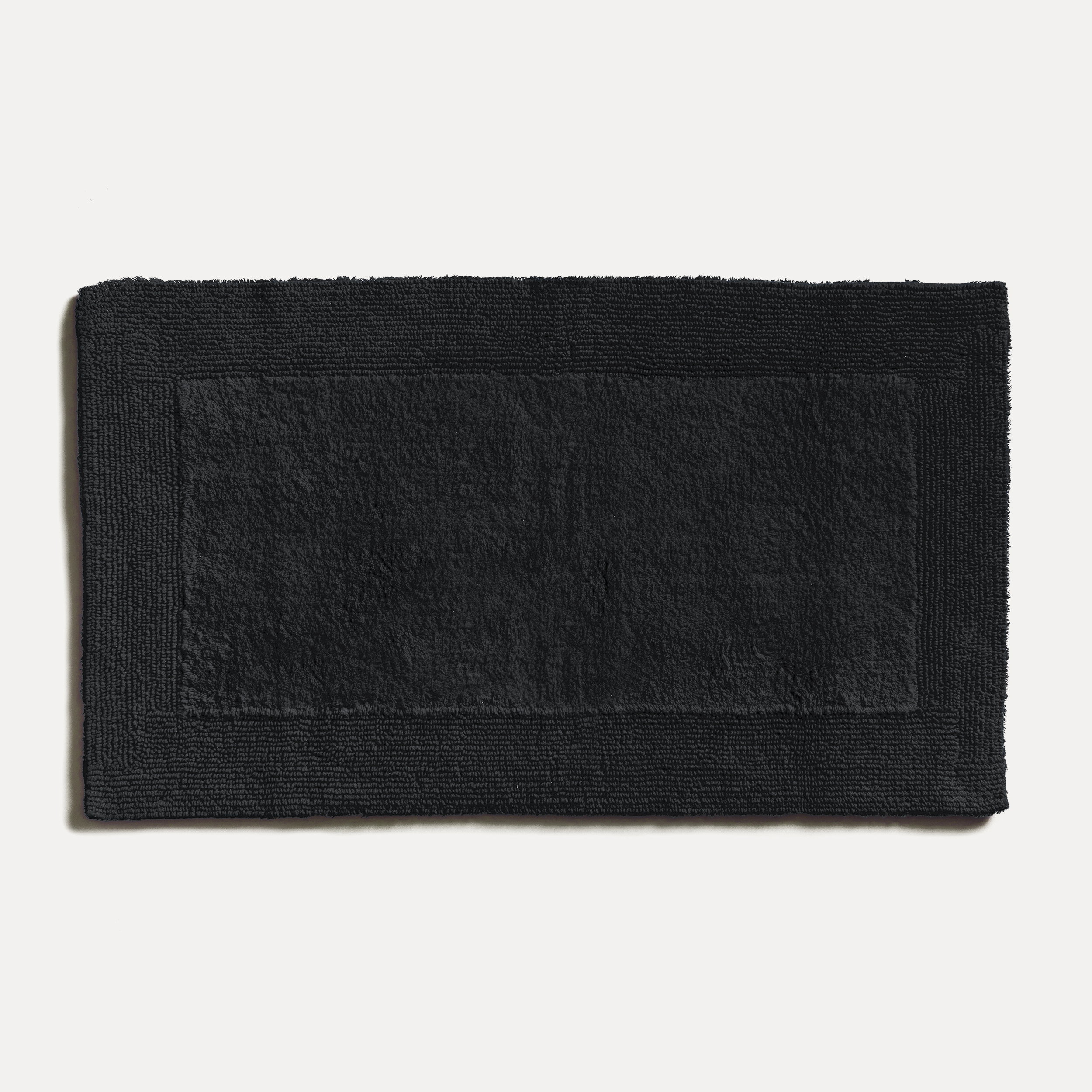 A black, rectangular bath mat lies flat on a light-colored surface; textured terry cloth surface with a subtle border, a simple, timeless bathroom accessory.