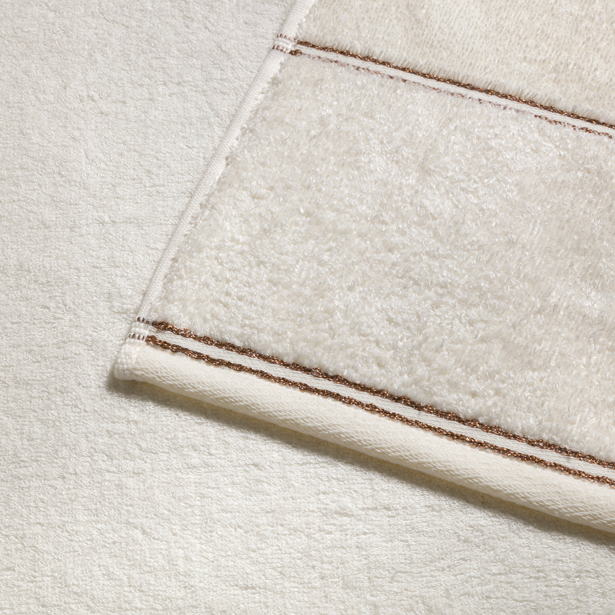 Cremefarbenes Frottierhandtuch mit feinem braunen Zierstreifen liegt gefaltet auf einem zweiten Handtuch; Nahaufnahme zeigt weiche Textur und Saumdetail.
