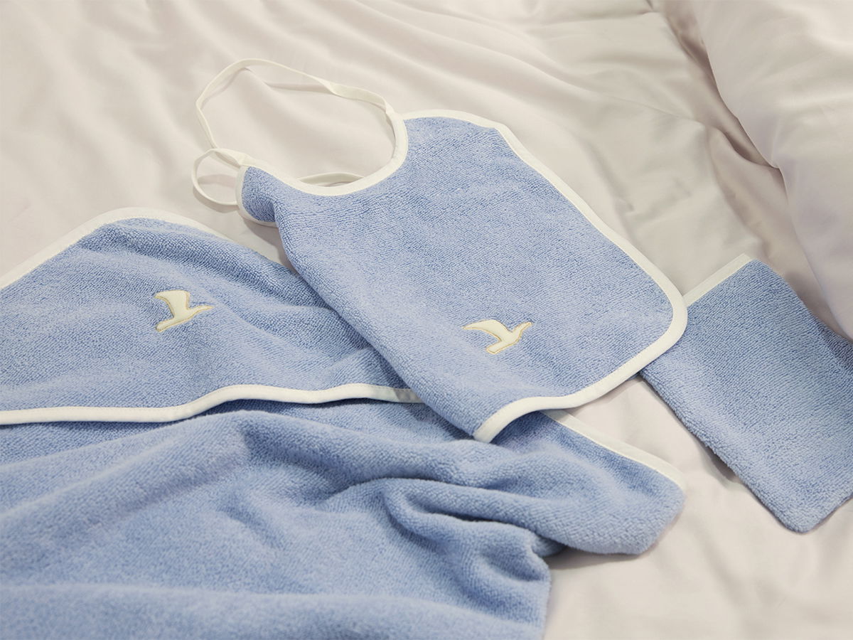 A light blue baby bib with a matching towel and washcloth are laid out on a light-colored bedsheet; soft terry cloth texture with white trim and a small seagull logo.