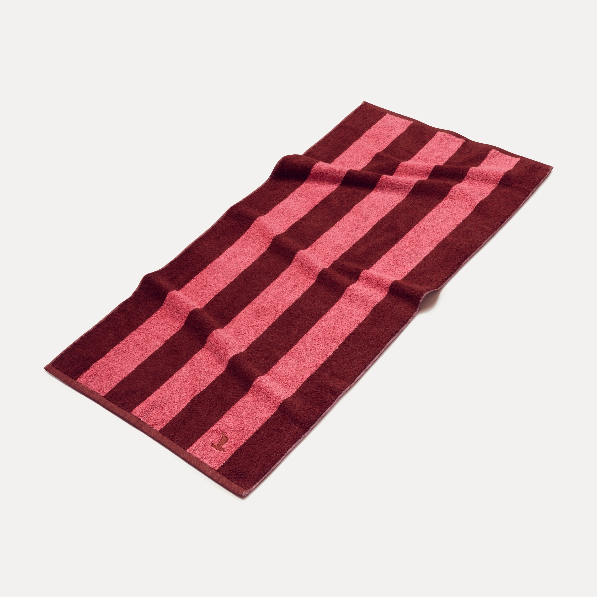 Gestreiftes Handtuch (dunkelrot und pink) liegt flach ausgebreitet mit kleinem Logo in der Ecke auf neutralem hellen Studiohintergrund.
