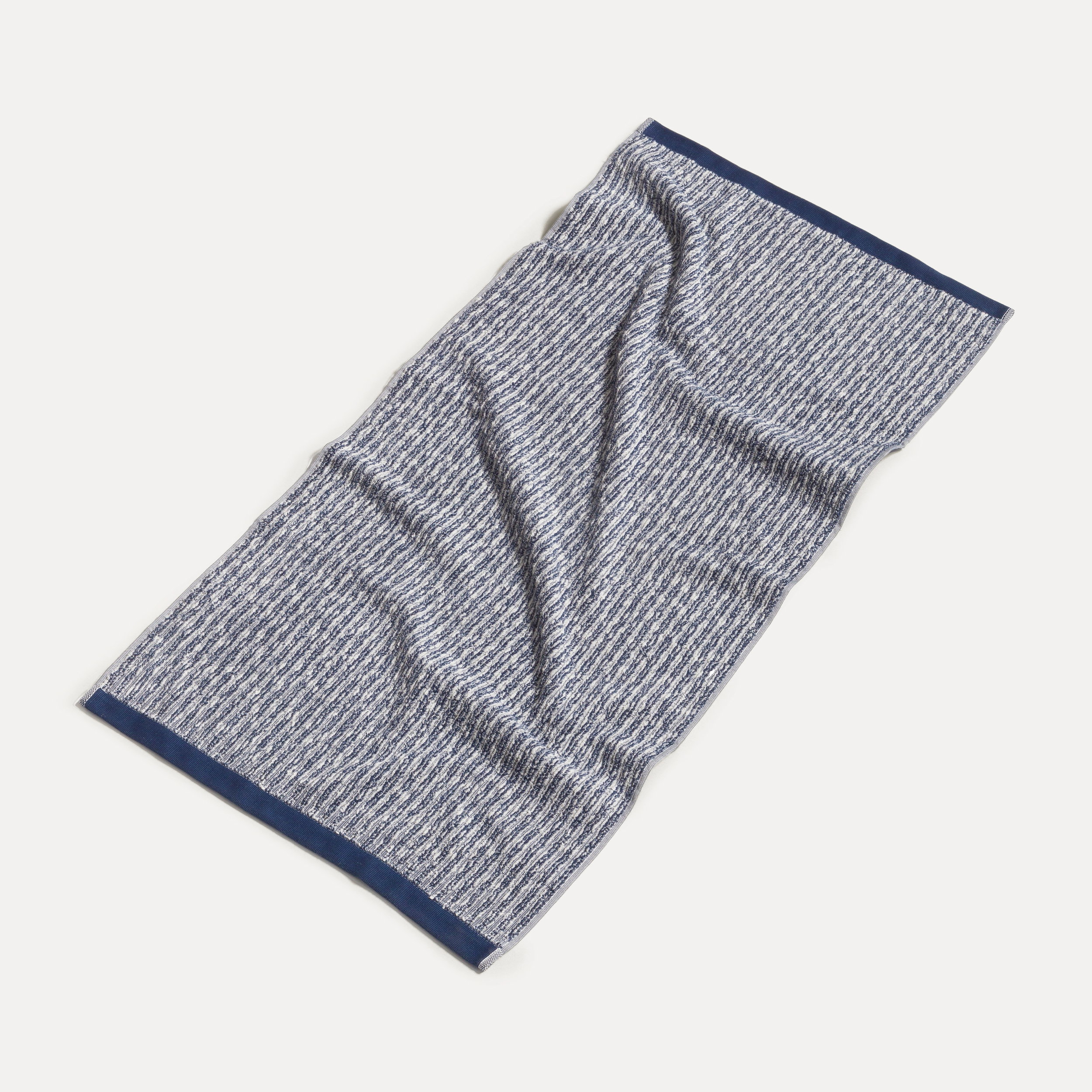 Handtuch liegt ausgebreitet und leicht zerknittert; fein strukturiertes blau-weißes Muster mit dunkelblauen Abschlusskanten, fotografiert auf hellem, neutralem Hintergrund.