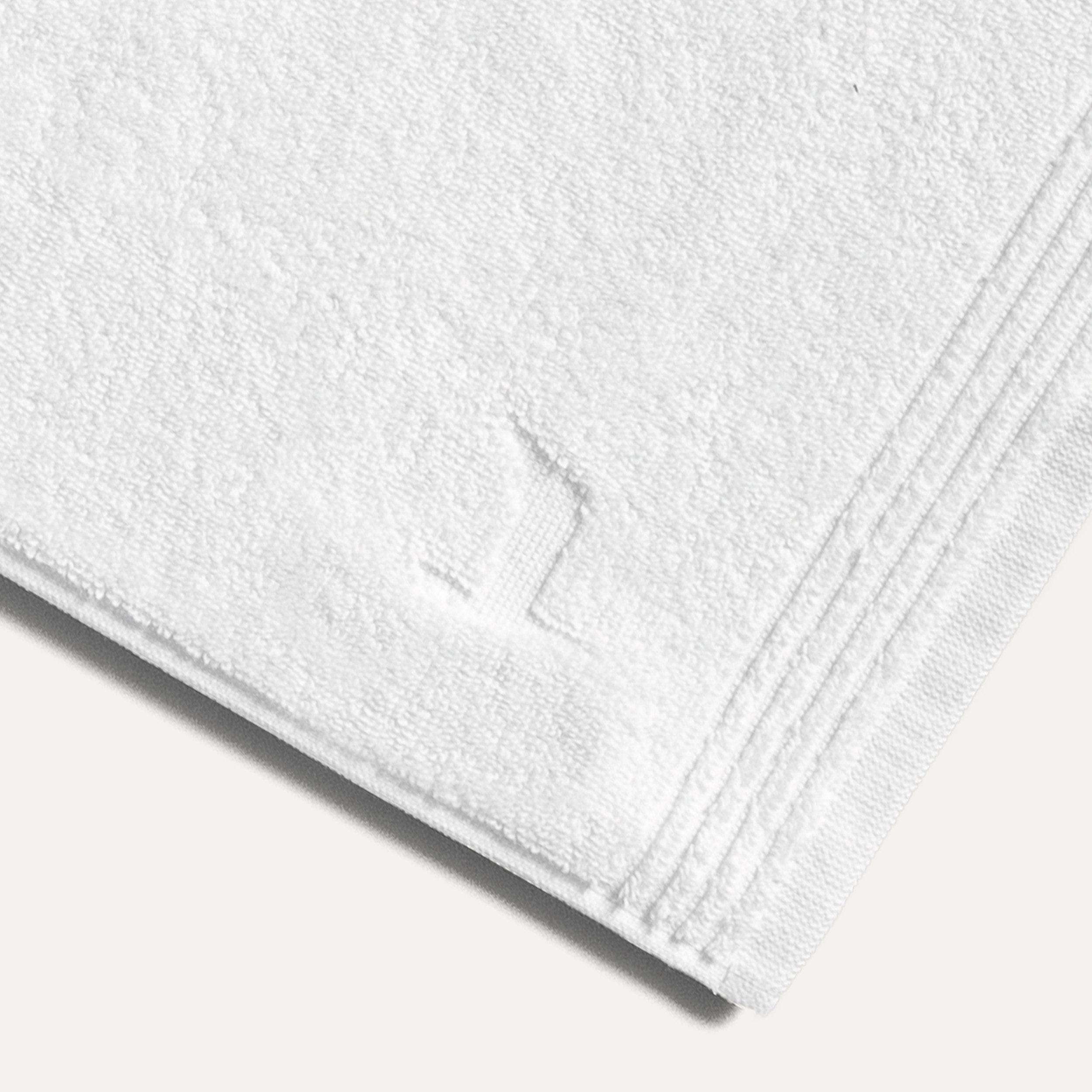 Weißes Frotteehandtuch, flach liegend mit eingeprägtem Monogramm und gefalteter Ecke, dekorative Ripsborden sichtbar; auf hellem, neutralem Untergrund.