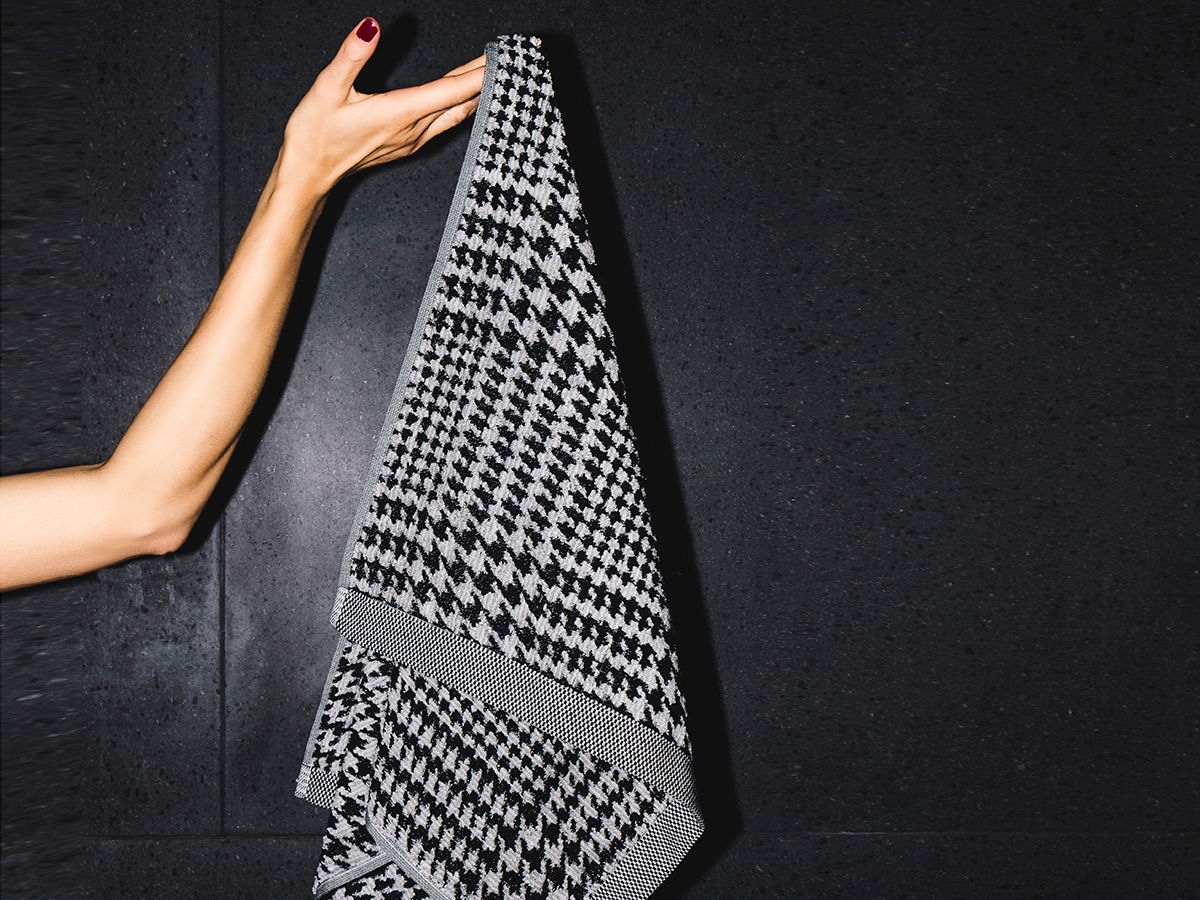 Handtuch — schwarz-weißes Hahnentrittmuster — wird von einer Hand an einer dunklen, strukturierten Wand gehalten; schlichtes Badezimmer-Accessoire, betont Kontrast und Textur.