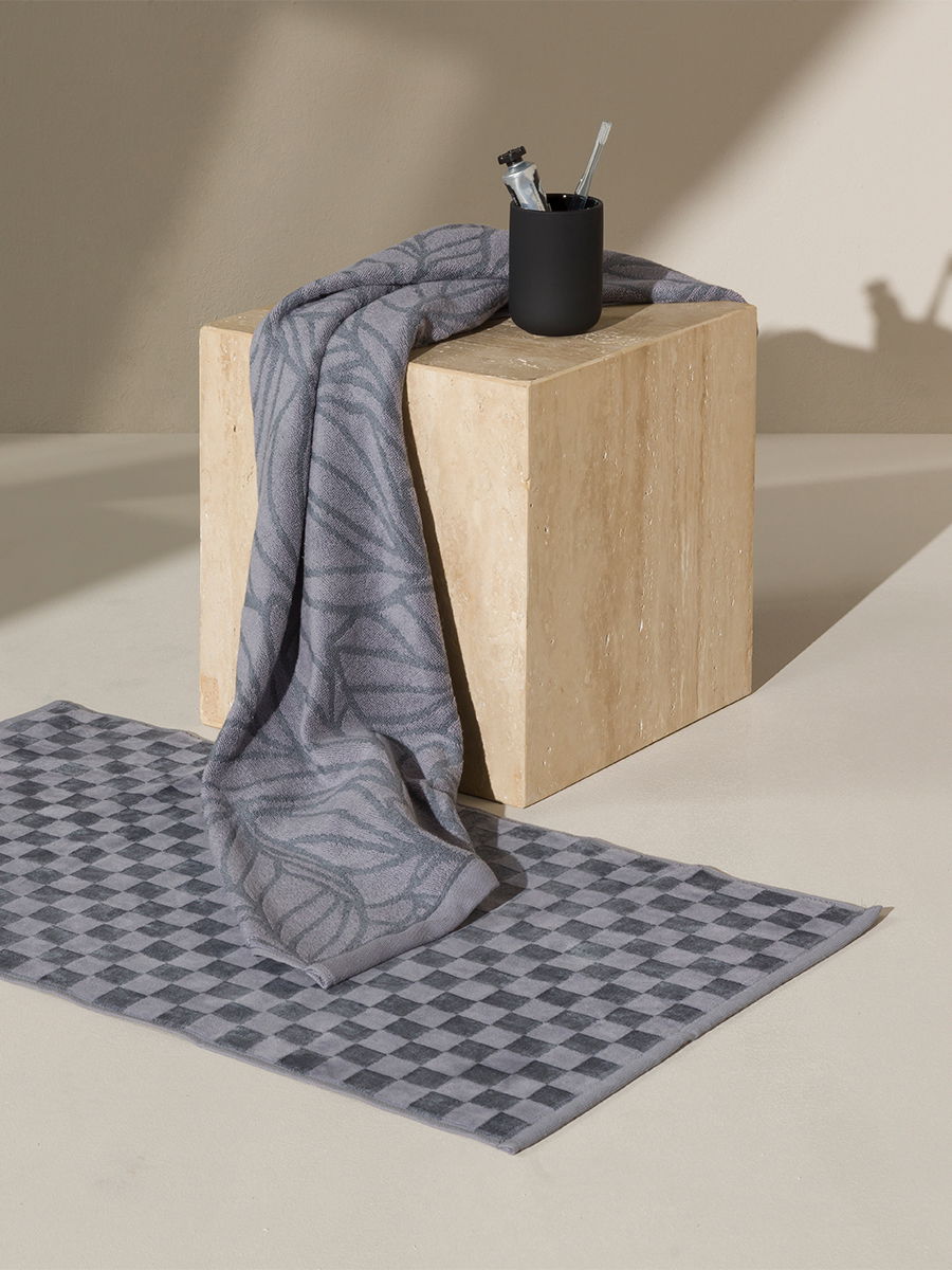 A gray, geometrically patterned towel lies across a light-colored, rectangular stone block and hangs down; in front of it is a checkered bath mat, next to it a black toothbrush cup with toothbrushes; neutral studio background.