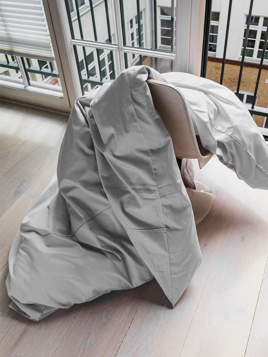 Weißes Textil (Bademantel oder großes Handtuch) drapiert über einem Stuhl und hängt bodenlang herunter; steht auf hellem Holzboden vor bodentiefen Fenstern mit Balkonbrüstung.