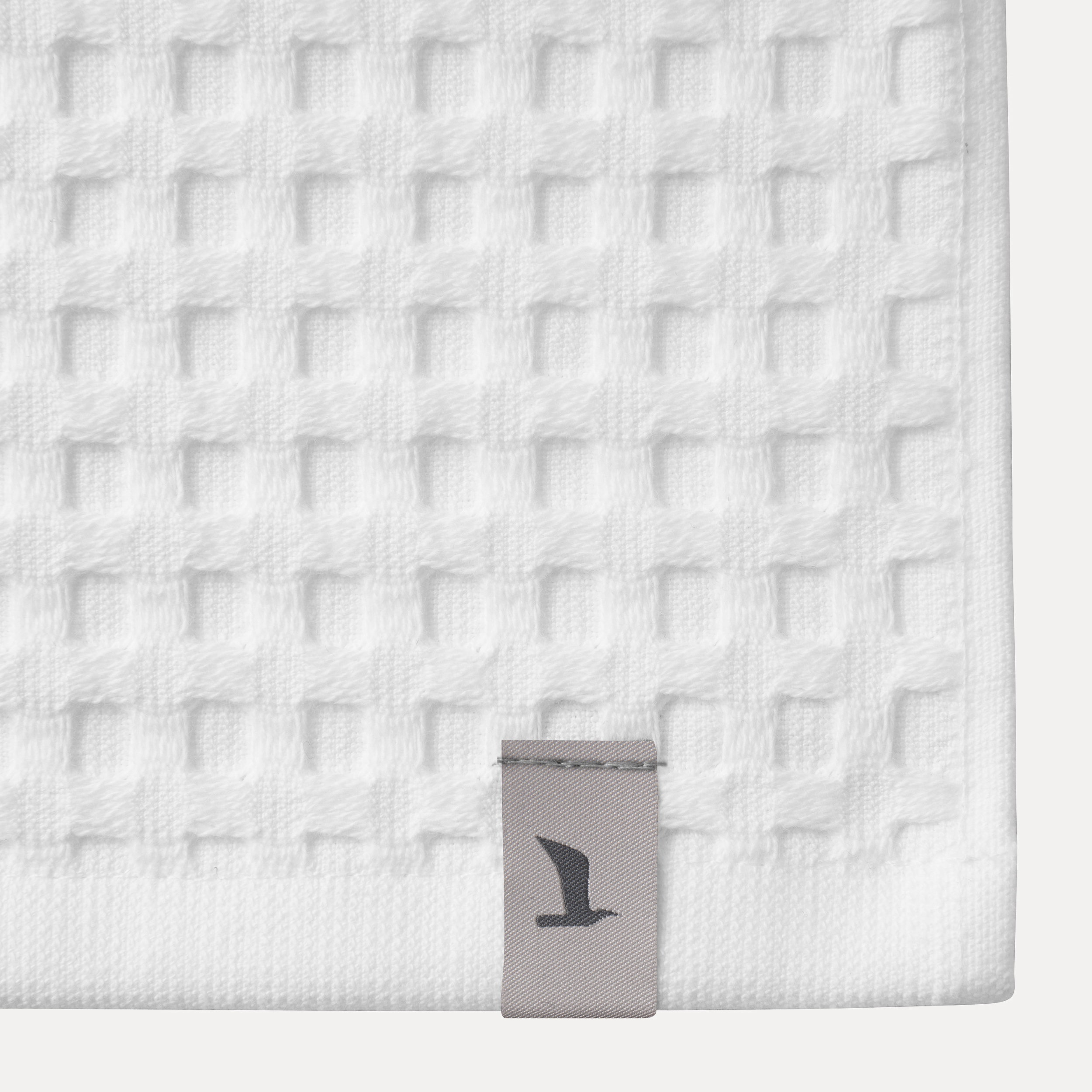 Towel — lying flat — white terry cloth with hem; gray label with seagull logo in the corner, light neutral background.