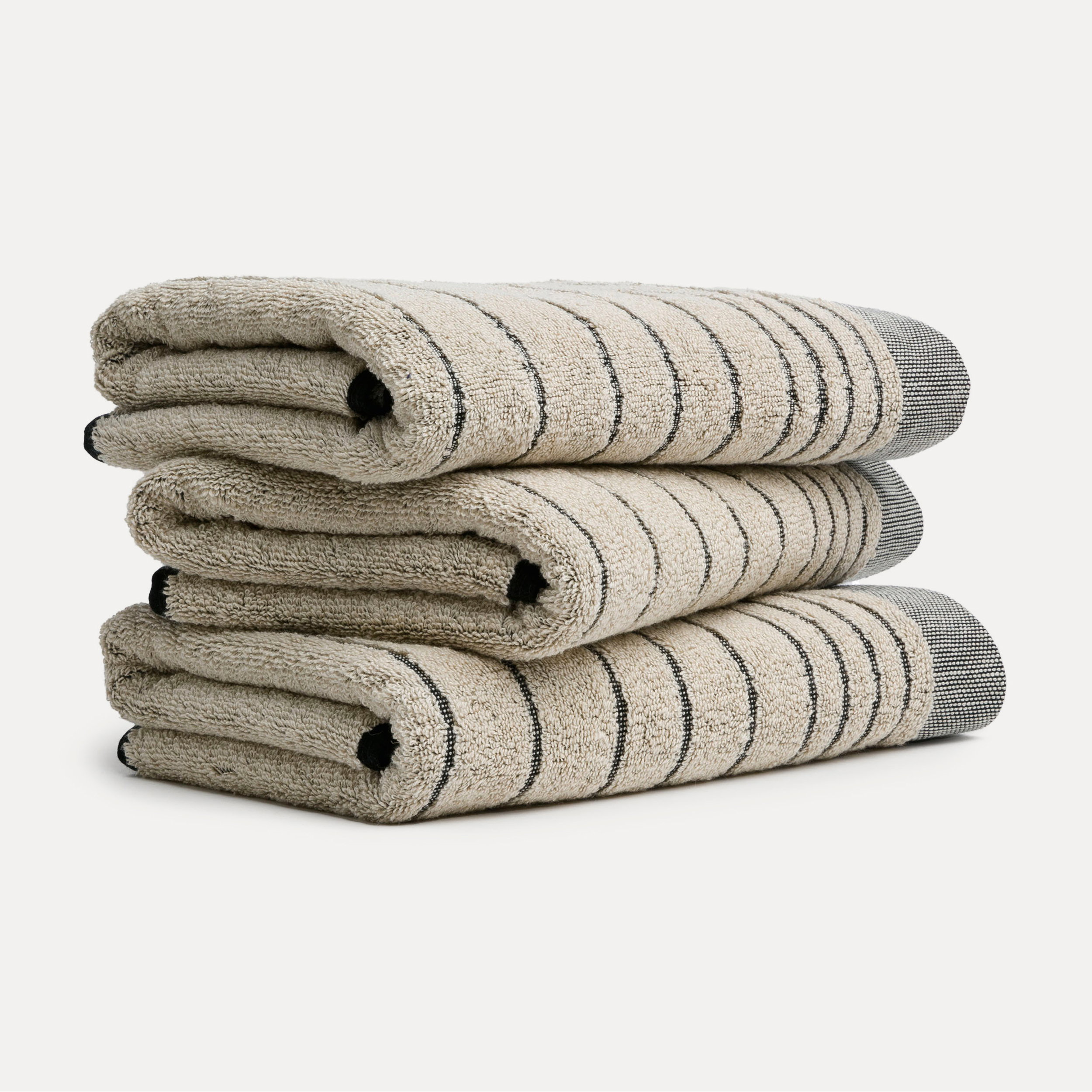 Three striped towels lie folded on top of one another; subtle beige-gray tones with dark gray edges, set against a neutral white background in a straightforward product shot.