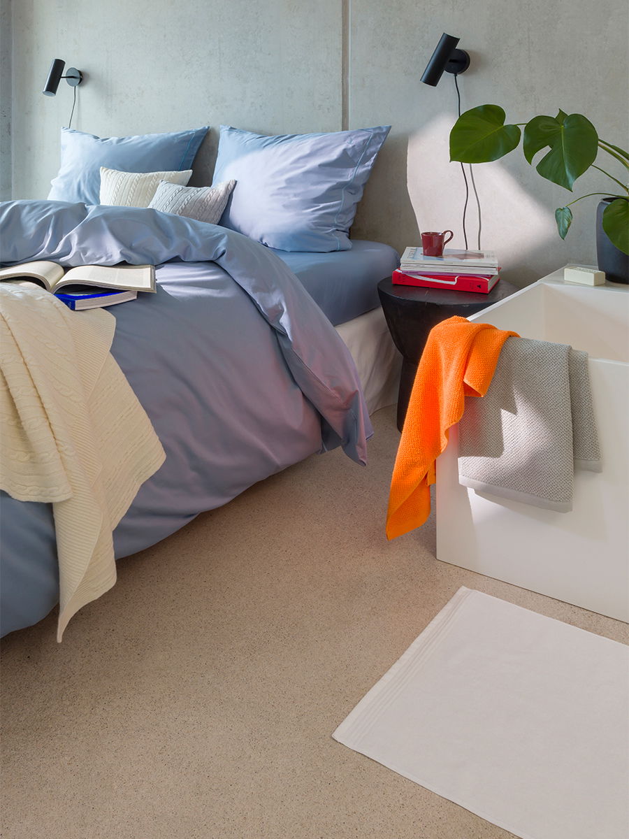 Oranges Handtuch hängt über einer Badewannenkante; weitere Handtücher und blaue Bettwäsche liegen auf dem Bett in einem hellen, kombinierten Schlaf- und Badezimmer mit Nachttisch und Pflanze.