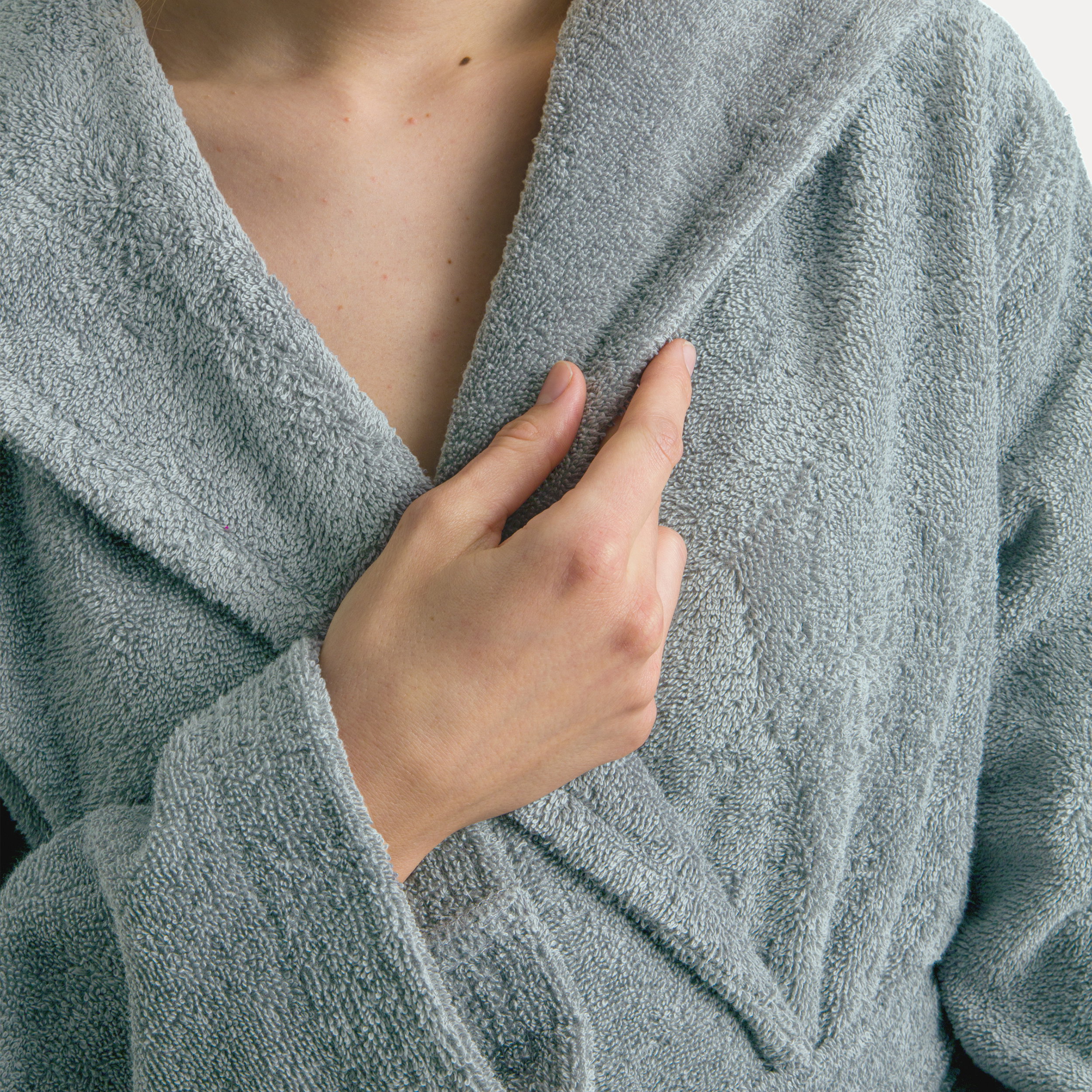 Terry cloth bathrobe in light mint gray; hand holding the lapel. Close-up of a person’s upper body, neutral background, soft, fluffy texture visible.
