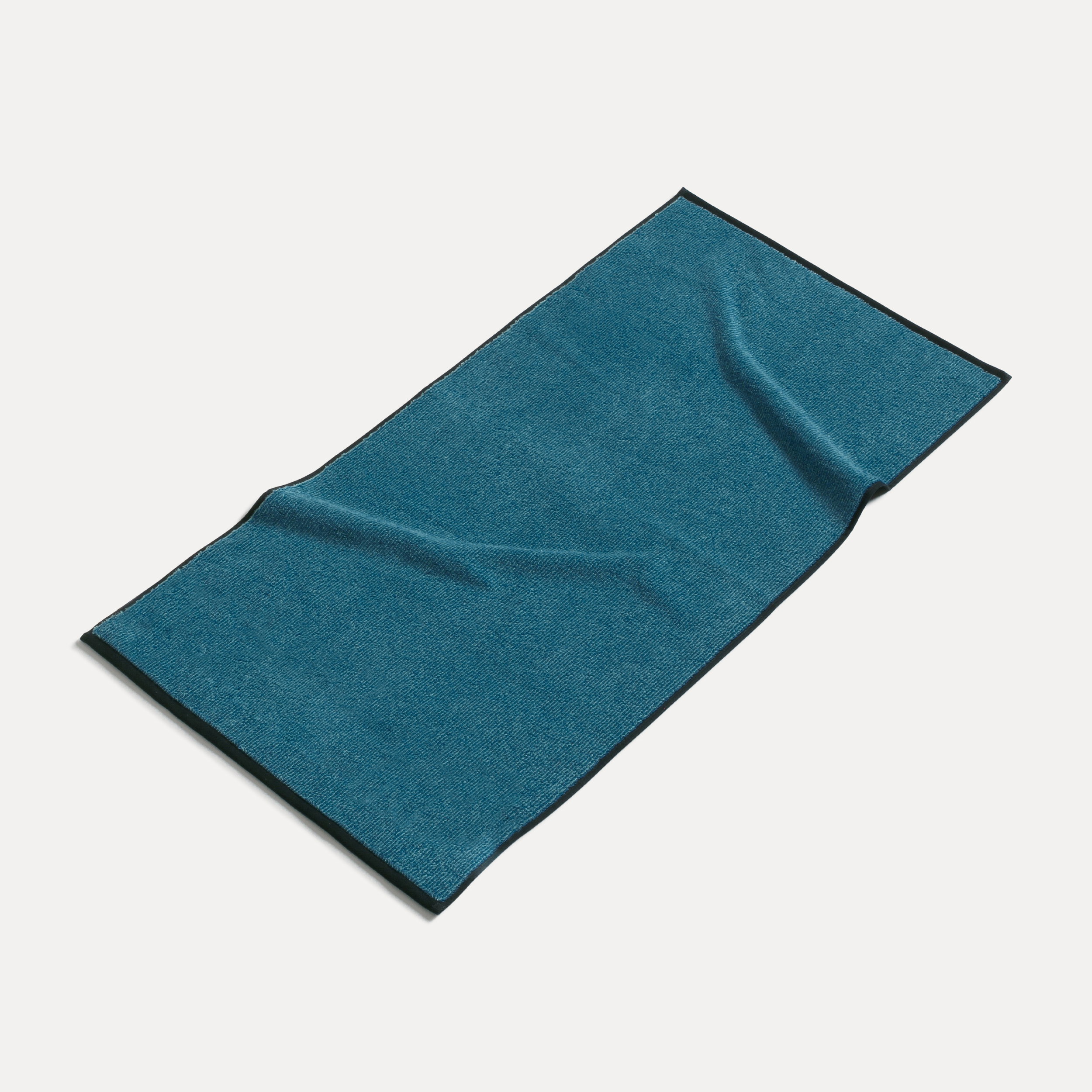 Handtuch liegt ausgebreitet auf neutralem, hellem Hintergrund; türkisblau mit dunkler Kante, leichte Falten, Stoffstruktur sichtbar, Studioaufnahme eines Badezimmer-Accessoires.