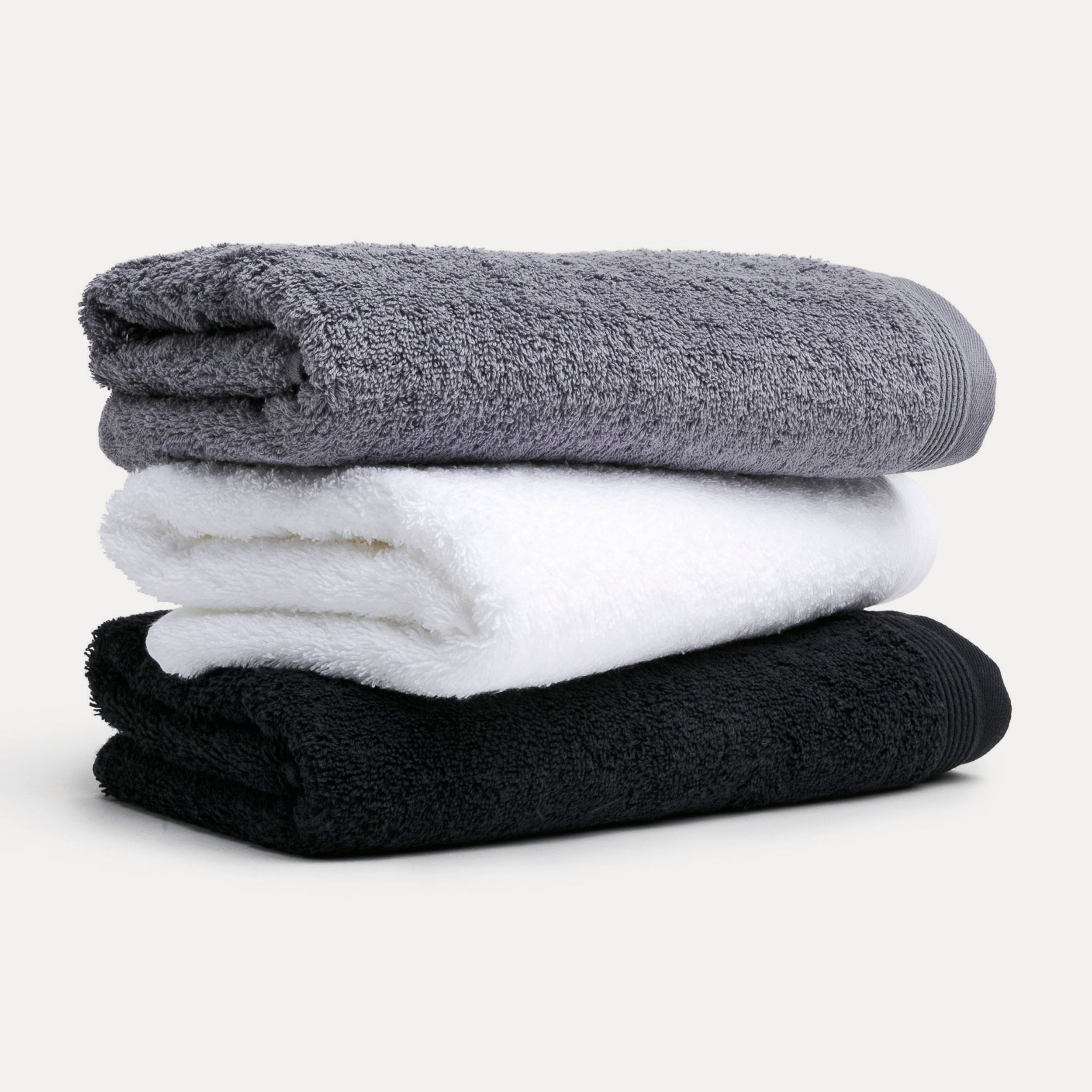 Three folded towels (gray, white, black) are stacked; they are neatly folded and display a fluffy texture against a neutral, light-colored studio background.