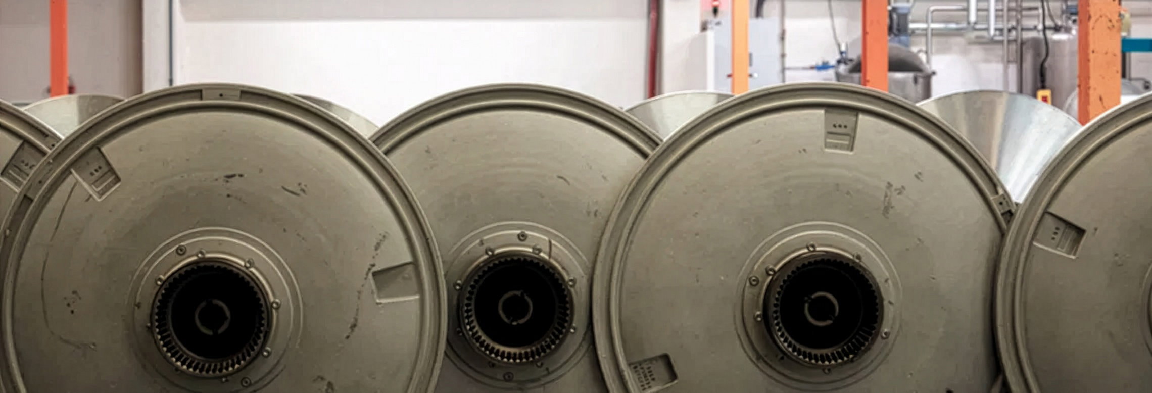 Mehrere große, runde Metalltrommeln stehen dicht nebeneinander; sichtbare Zahnringe in ihren Mittelpunkten, in einer hellen Industriehalle mit Stahlrohren und orangefarbenen Stützen.