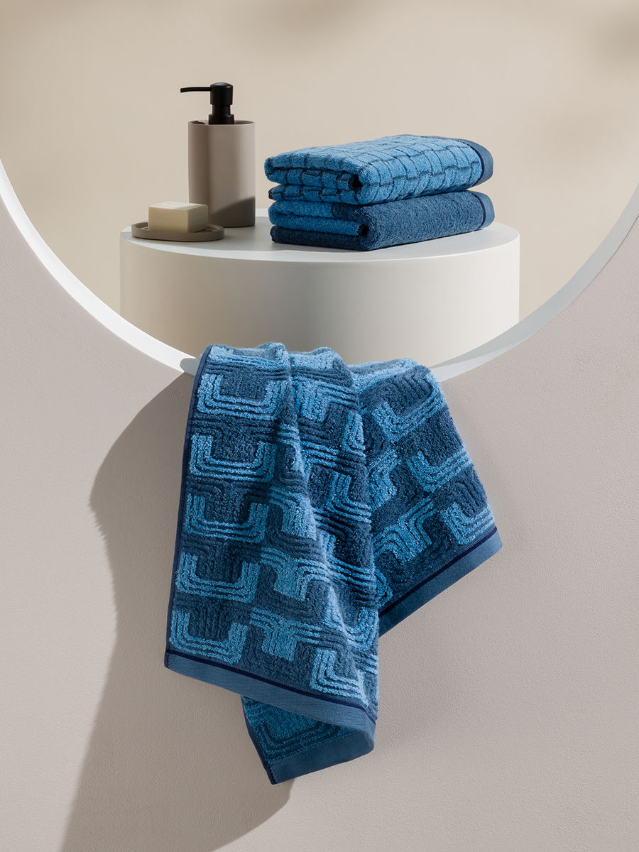 Blue, geometrically patterned towels: one hangs down, two lie folded on a round column; next to them, a soap dispenser and bar of soap in a neutral, minimalist bathroom setting.