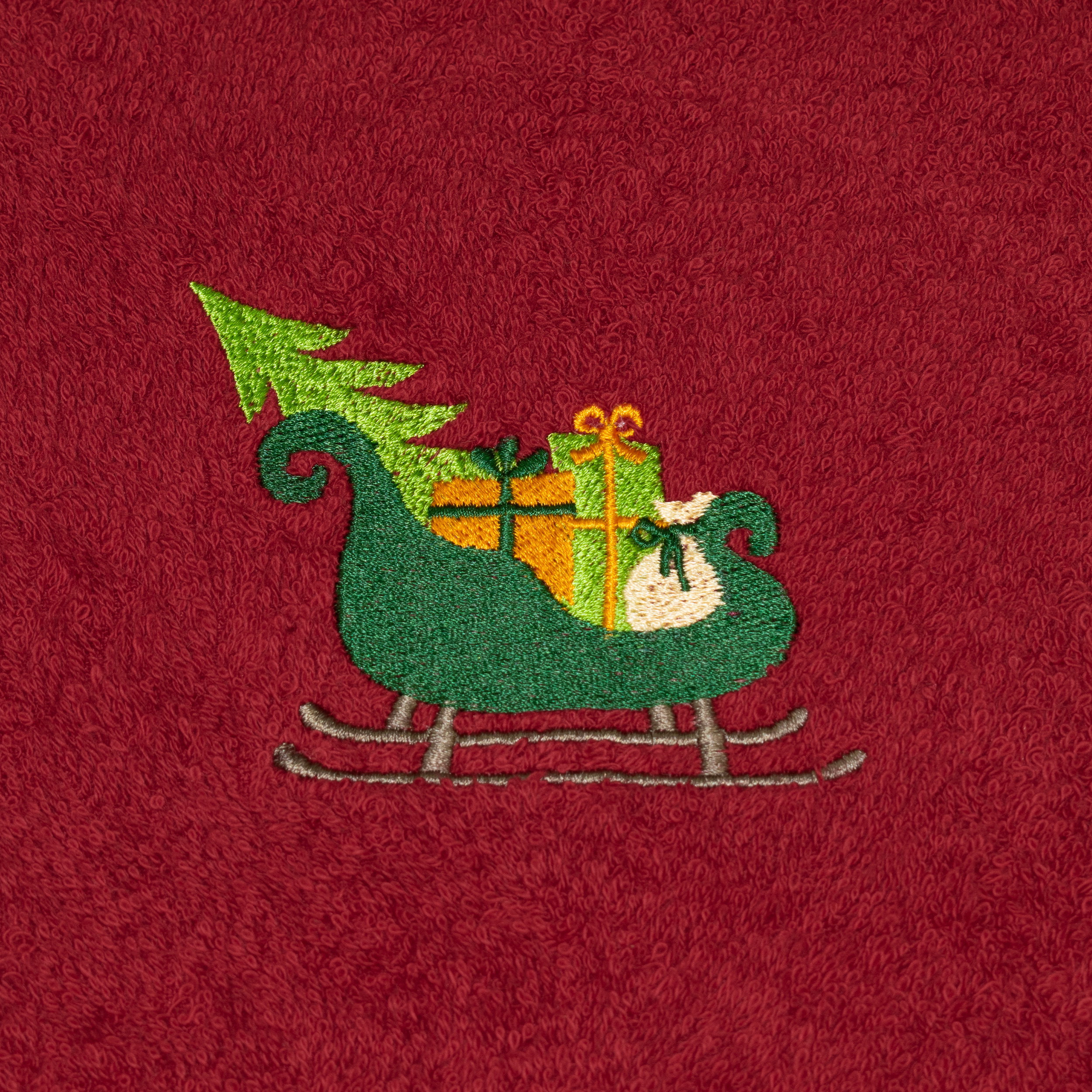 Rotes Frotteehandtuch zeigt gestickten grünen Schlitten, der Geschenke und einen Tannenbaum trägt; Nahaufnahme auf roter Frotteefläche.