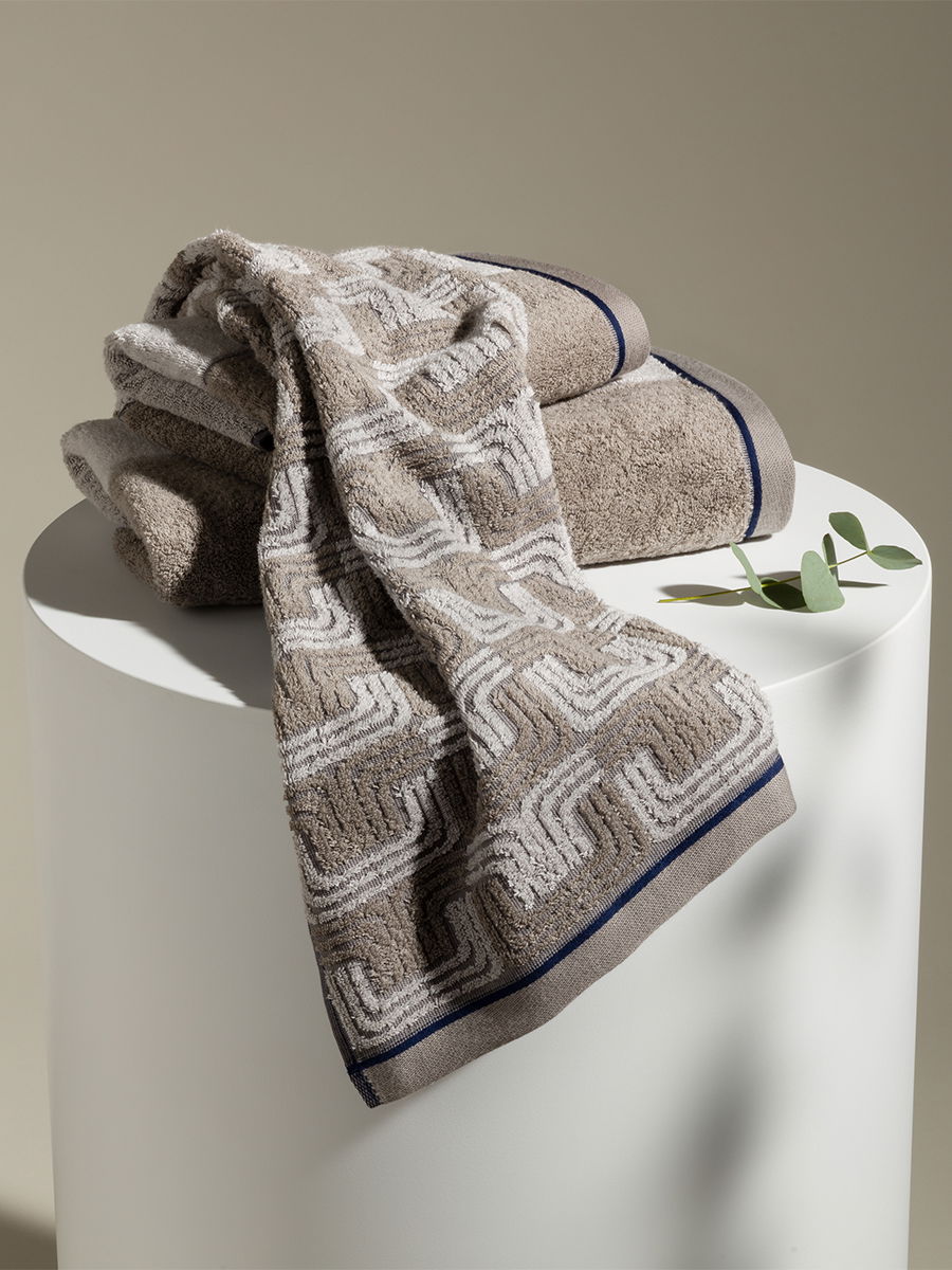 Towels are stacked on a round white pedestal; one draped down at the front shows a woven pattern. Simple, neutral colors; subtle plant branch and light background.