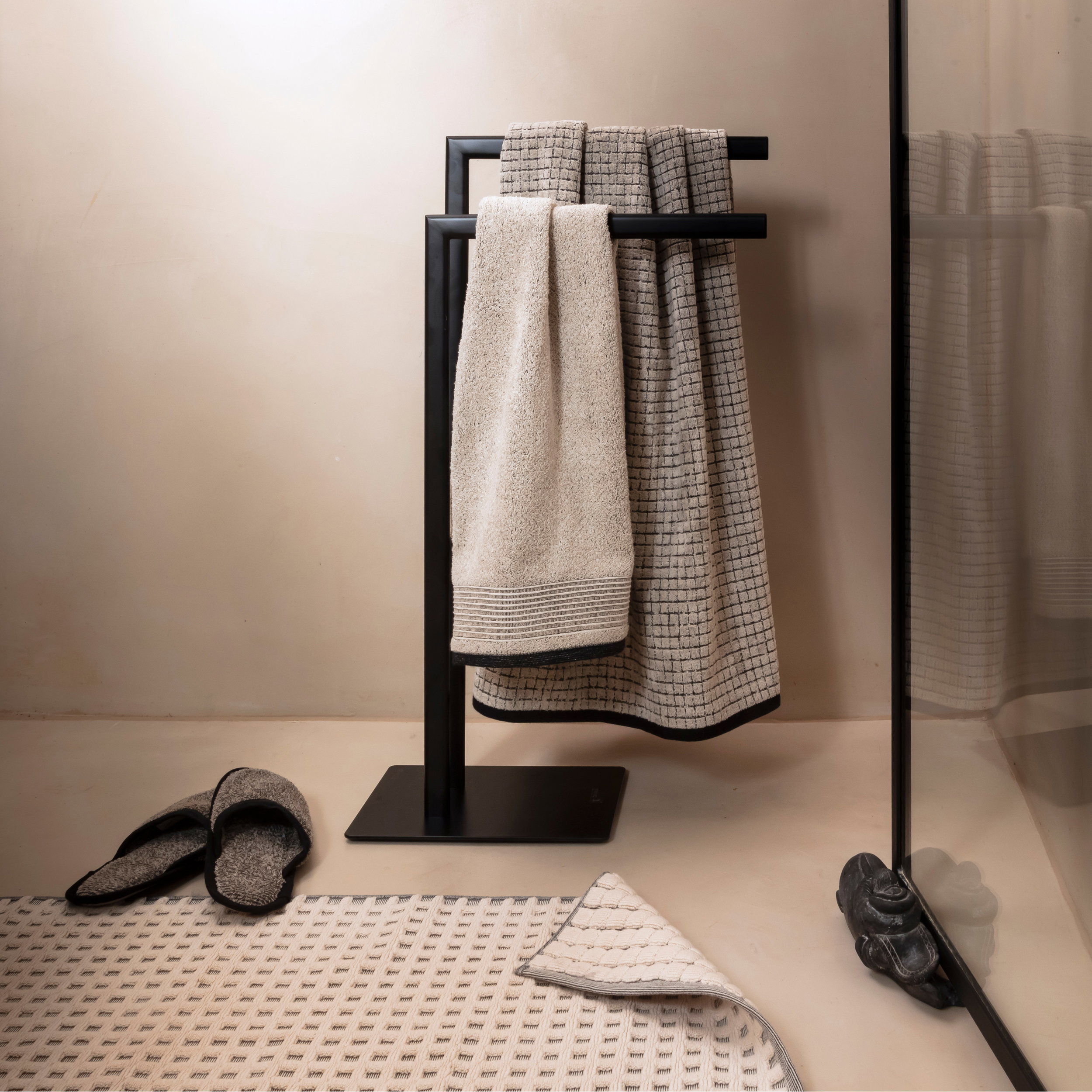 Zwei strukturierte Handtücher hängen an einem schwarzen, freistehenden Handtuchhalter; daneben Badmatte, Hausschuhe und eine Glas-Duschabtrennung in einem hellbeigen, minimalistischen Badezimmer.
