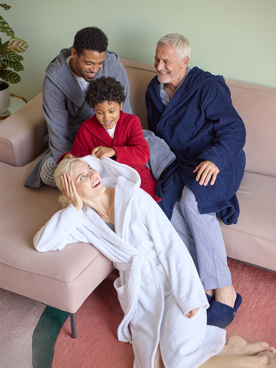Vier Personen tragen unterschiedliche Bademäntel; sie sitzen und liegen entspannt auf einem Sofa, lachen und interagieren in einer hellen, wohnzimmerähnlichen Umgebung mit Teppich und Pflanzen.