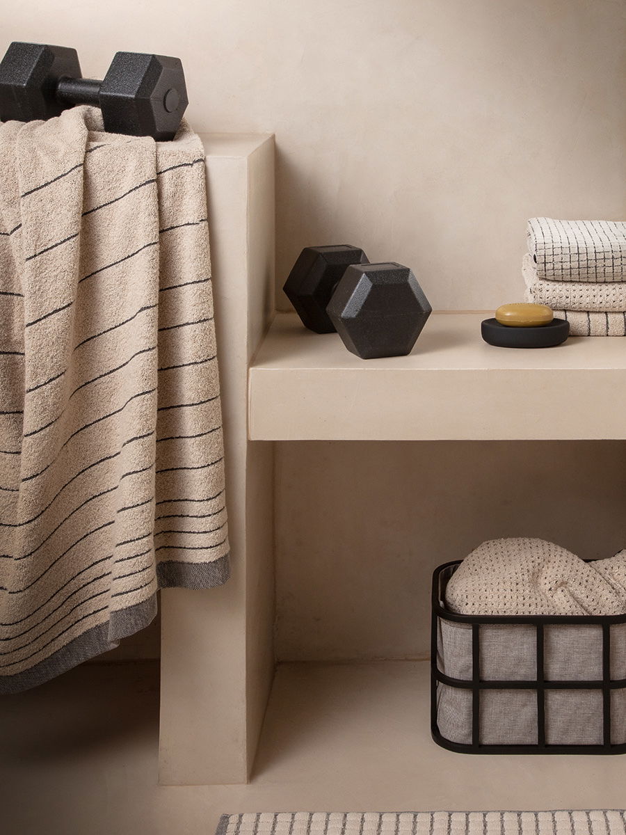 Gestreiftes Handtuch hängt über einer beige Sitzbank; daneben zwei Kurzhanteln, Seifenschale, gefaltete Handtücher und ein Drahtkorb mit Textil in einem minimalistischen Badezimmer.
