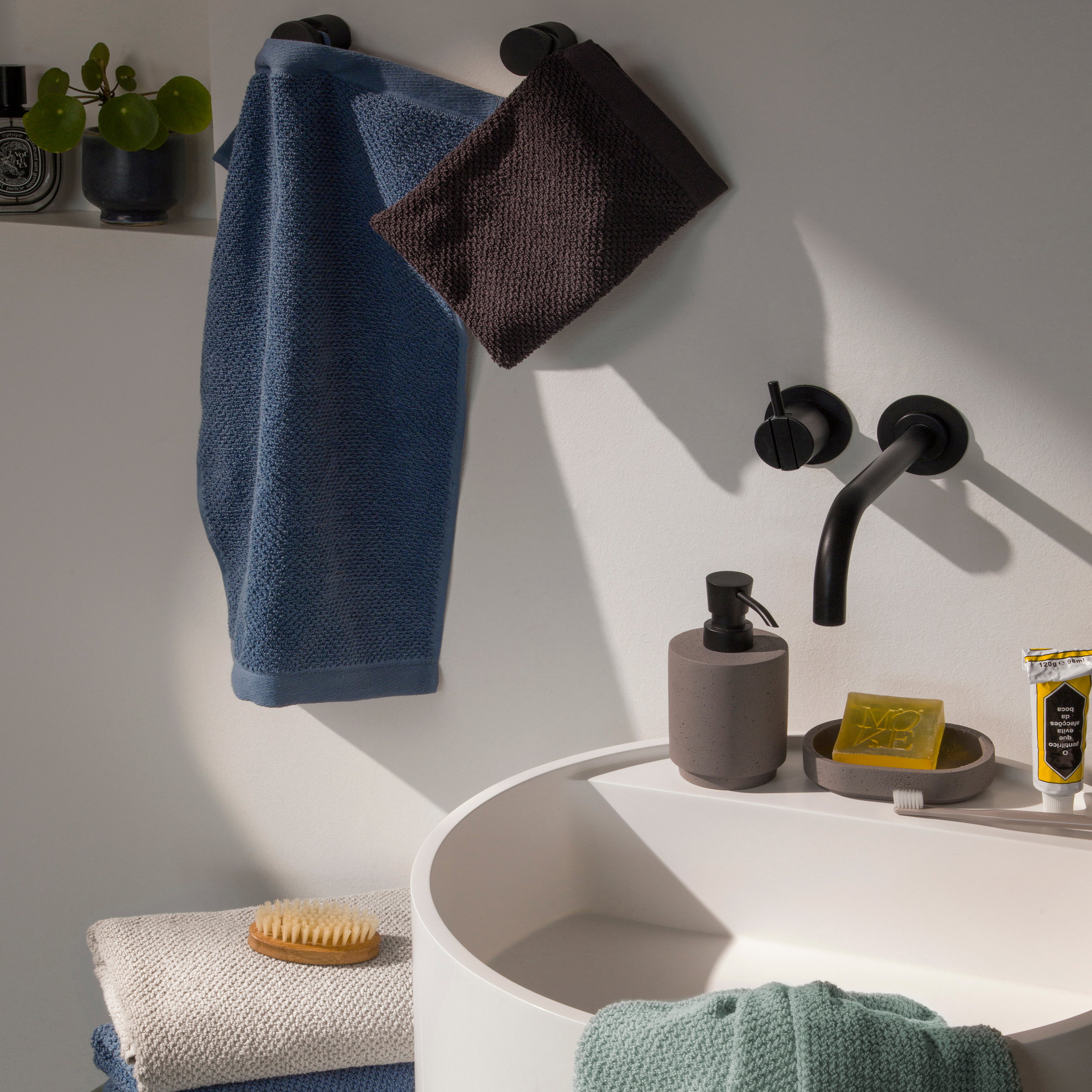 Blue and dark gray towels hang on wall hooks, complemented by folded towels and a brush; modern bathroom with a white round washbasin, black wall-mounted faucet, soap dispenser, and soap dish.