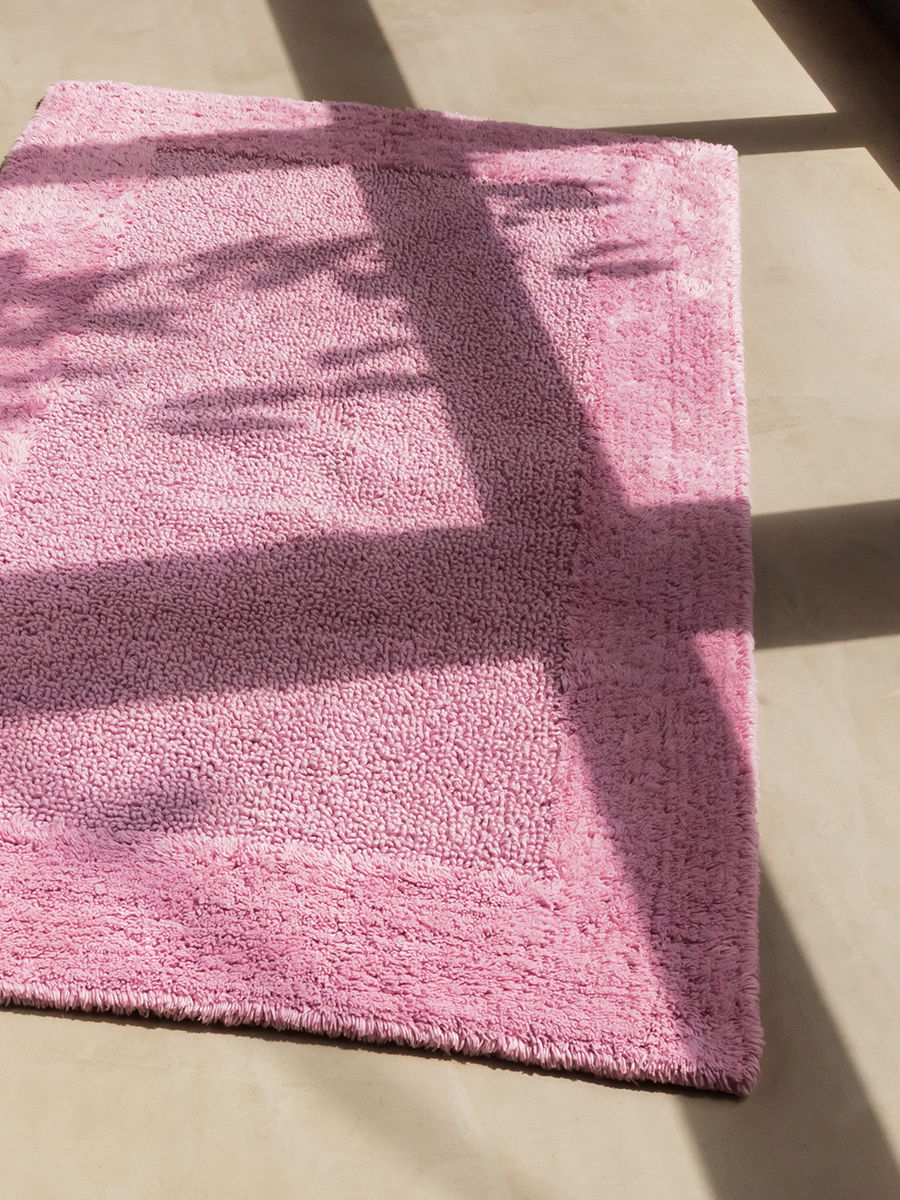 Ein rosa Badvorleger liegt auf hellem Badezimmerboden, von Fensterrahmen- und Pflanzenschatten durchzogen und von warmem Sonnenlicht beleuchtet.